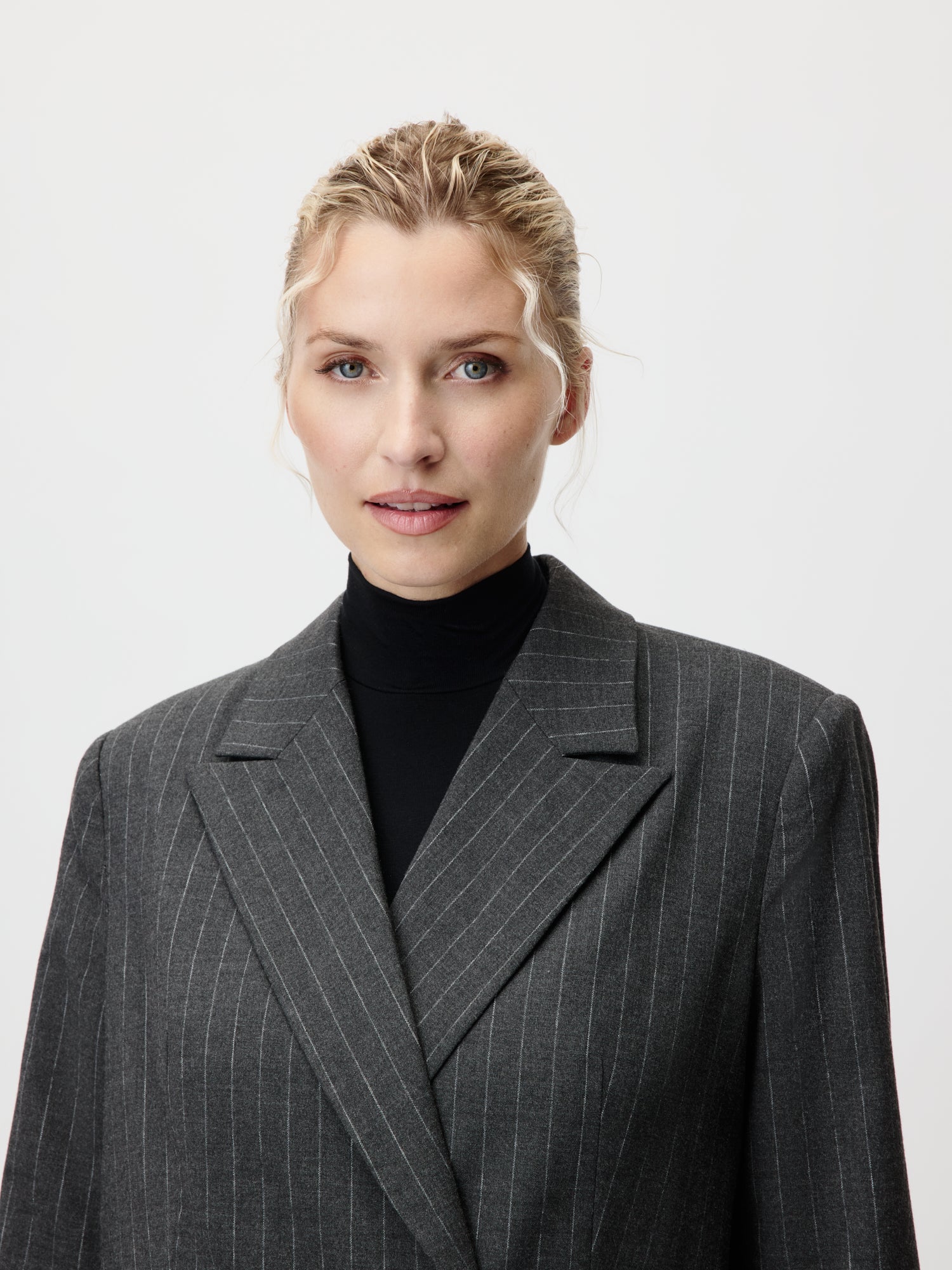 Eine selbstbewusste Frau mit zurückgebundenen blonden Haaren trägt einen dunkelgrauen Nadelstreifen-Blazer Delphine von LeGer by Lena Gercke in Größe 36 über einem schwarzen Rollkragenpullover, steht vor einem schlichten hellen Hintergrund und blickt in die Kamera.