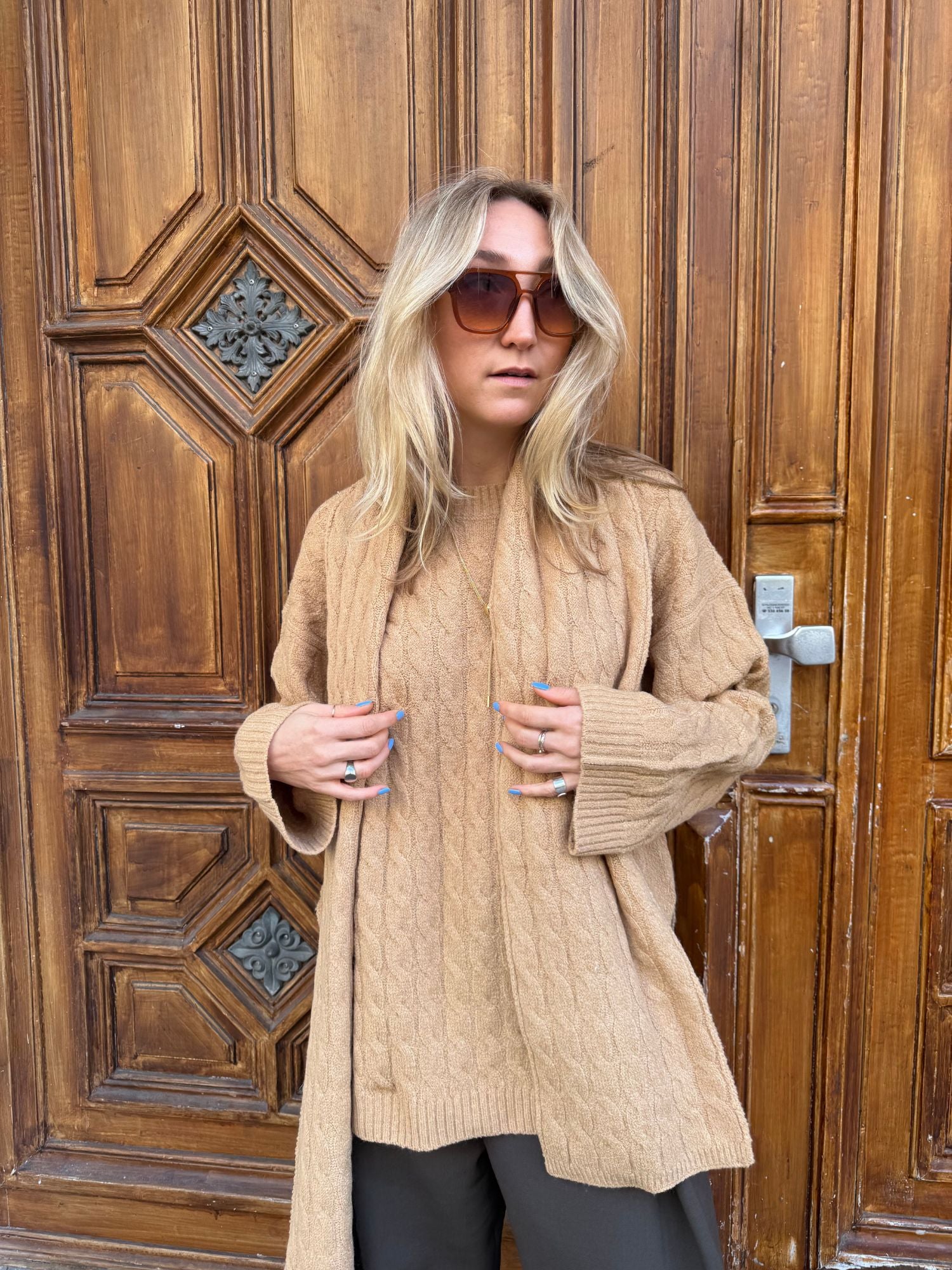 Eine Frau mit langen blonden Haaren, einer übergroßen Sonnenbrille und dem Jamila Jumper von LeGer by Lena Gercke in Größe 36 und langer Passform steht vor einer großen, kunstvoll getäfelten Holztür.