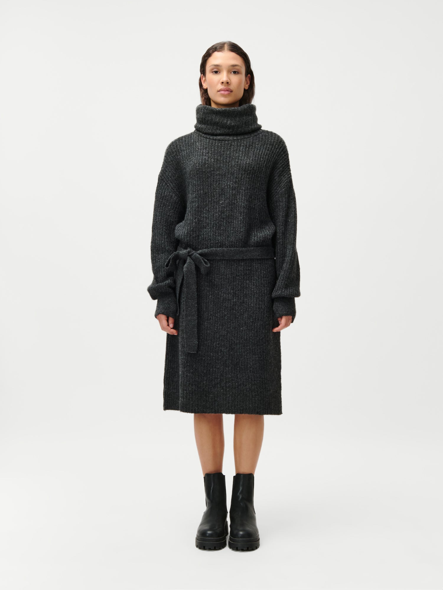 Eine Person blickt nach vorne und trägt das knielange, dunkelgraue Langarm-Rollkragenkleid „Colleen“ mit Taillengürtel von LeGer by Lena Gercke sowie schwarze Stiefeletten vor weißem Hintergrund.