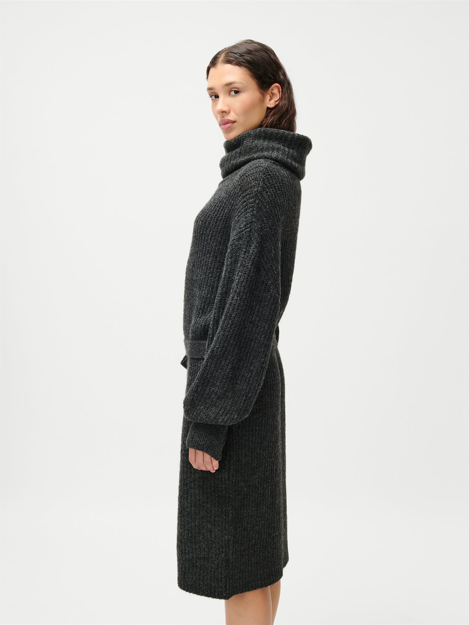 Eine Frau mit welligem braunem Haar trägt das knielange, dunkelgraue, gerippte Strickkleid „Colleen“ mit langen Ärmeln, normaler Passform und Taschen von LeGer by Lena Gercke und steht seitlich vor einem schlichten weißen Hintergrund.