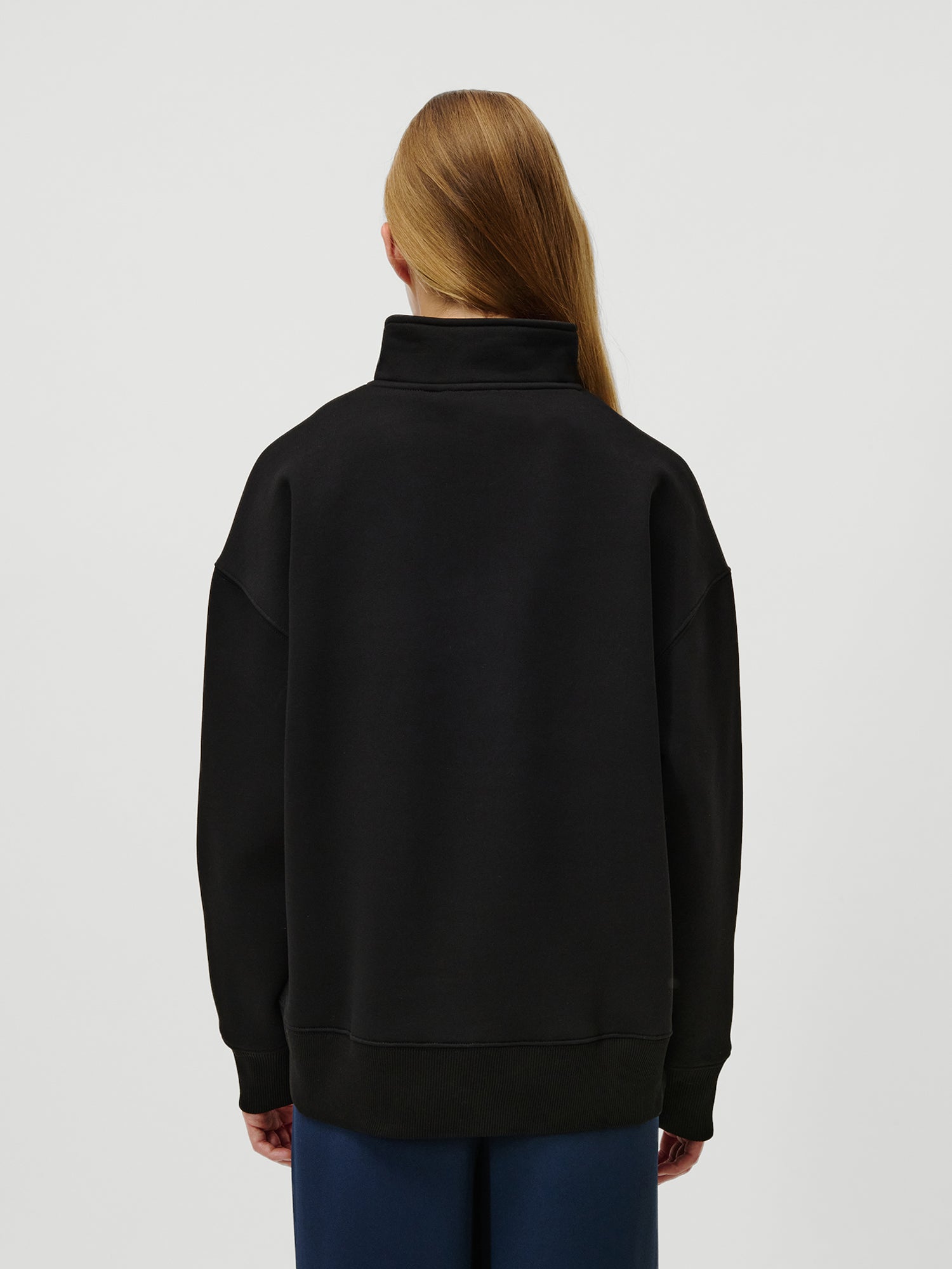 Eine Person mit langen, glatten, hellbraunen Haaren ist von hinten zu sehen. Sie trägt den Jenny Sweater von LeGer by Lena Gercke in Schwarz und eine marineblaue Hose vor einem schlichten, hellen Hintergrund.