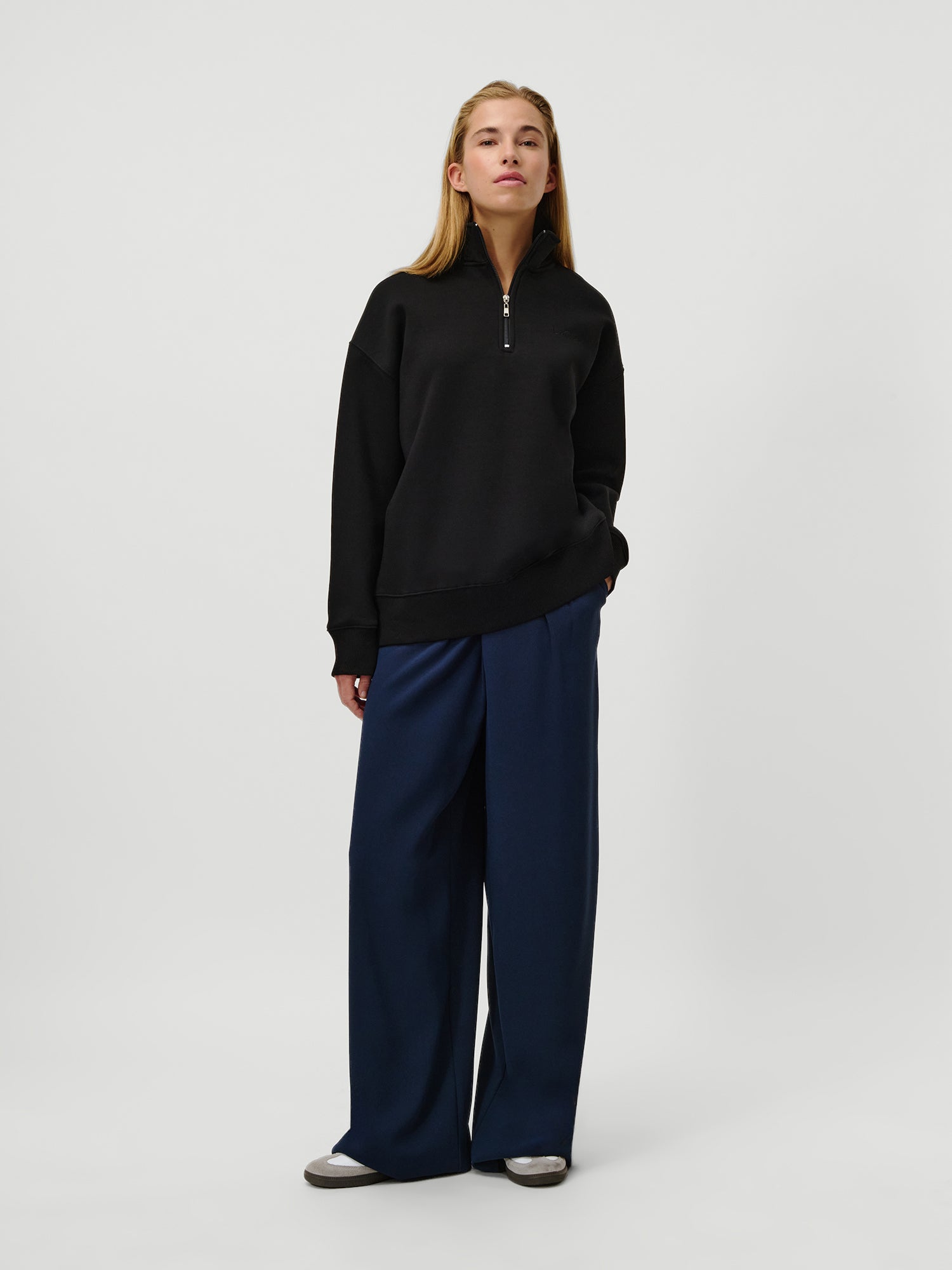 Eine Person mit langen blonden Haaren und einem neutralen Gesichtsausdruck steht vor einem weißen Hintergrund. Sie trägt den LeGer by Lena Gercke Jenny Sweater in Schwarz mit langen Ärmeln, eine marineblaue Hose mit weitem Bein und graue Schuhe und hat eine Hand in der Tasche.