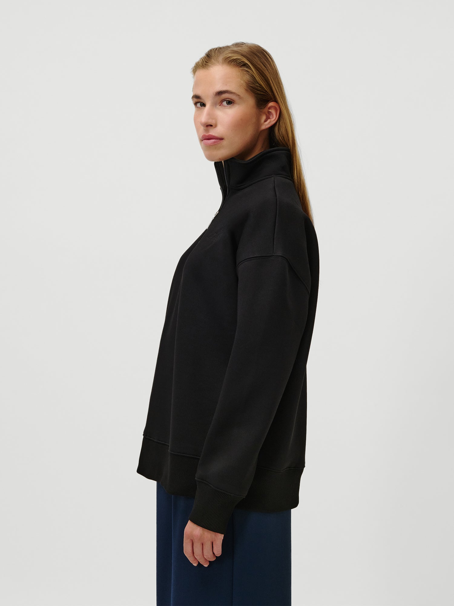 Eine Person mit langem, hellbraunem Haar trägt den LeGer by Lena Gercke Jenny Sweater - schwarz, locker sitzend, langärmelig, reguläre Länge - gepaart mit einer dunklen Hose und steht im Profil vor einem weißen Hintergrund.