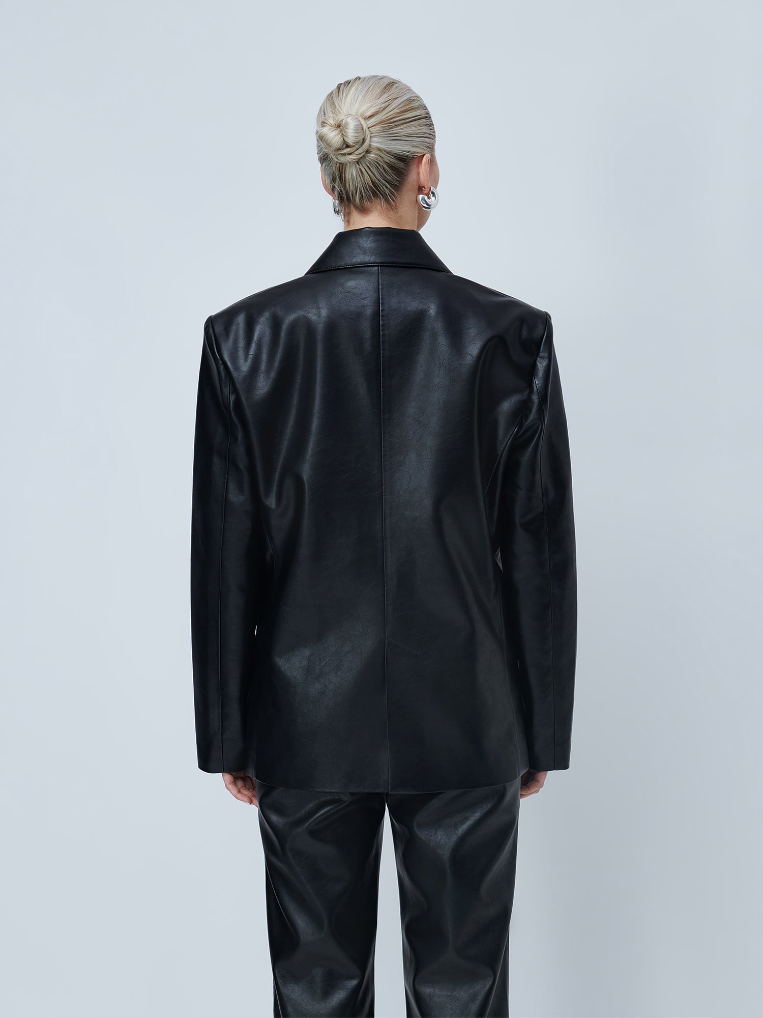 Eine Person mit blonden Haaren, die zu einem ordentlichen Dutt zusammengebunden sind, ist von hinten zu sehen. Sie trägt den schwarzen, einreihigen „Insa“-Blazer von LeGer by Lena Gercke in taillierter Passform sowie eine dazu passende schwarze Lederhose vor einem hellgrauen Hintergrund.