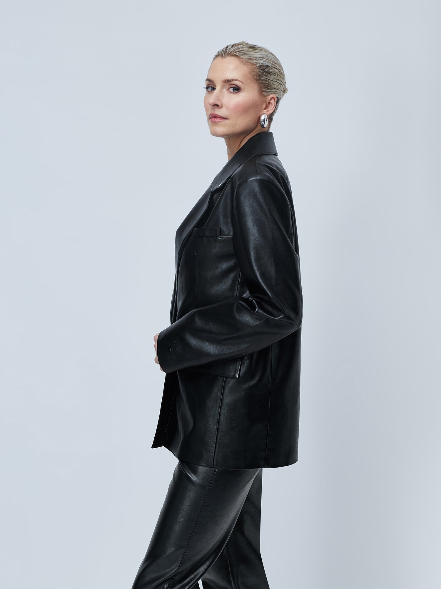 Eine Person mit blondem, nach hinten geglättetem Haar trägt den Blazer „Insa“ von LeGer by Lena Gercke in einreihiger, taillierter Passform mit passender Hose und posiert seitlich vor einem hellgrauen Hintergrund. Silberne Statement-Ohrringe unterstreichen ihren selbstbewussten Look.