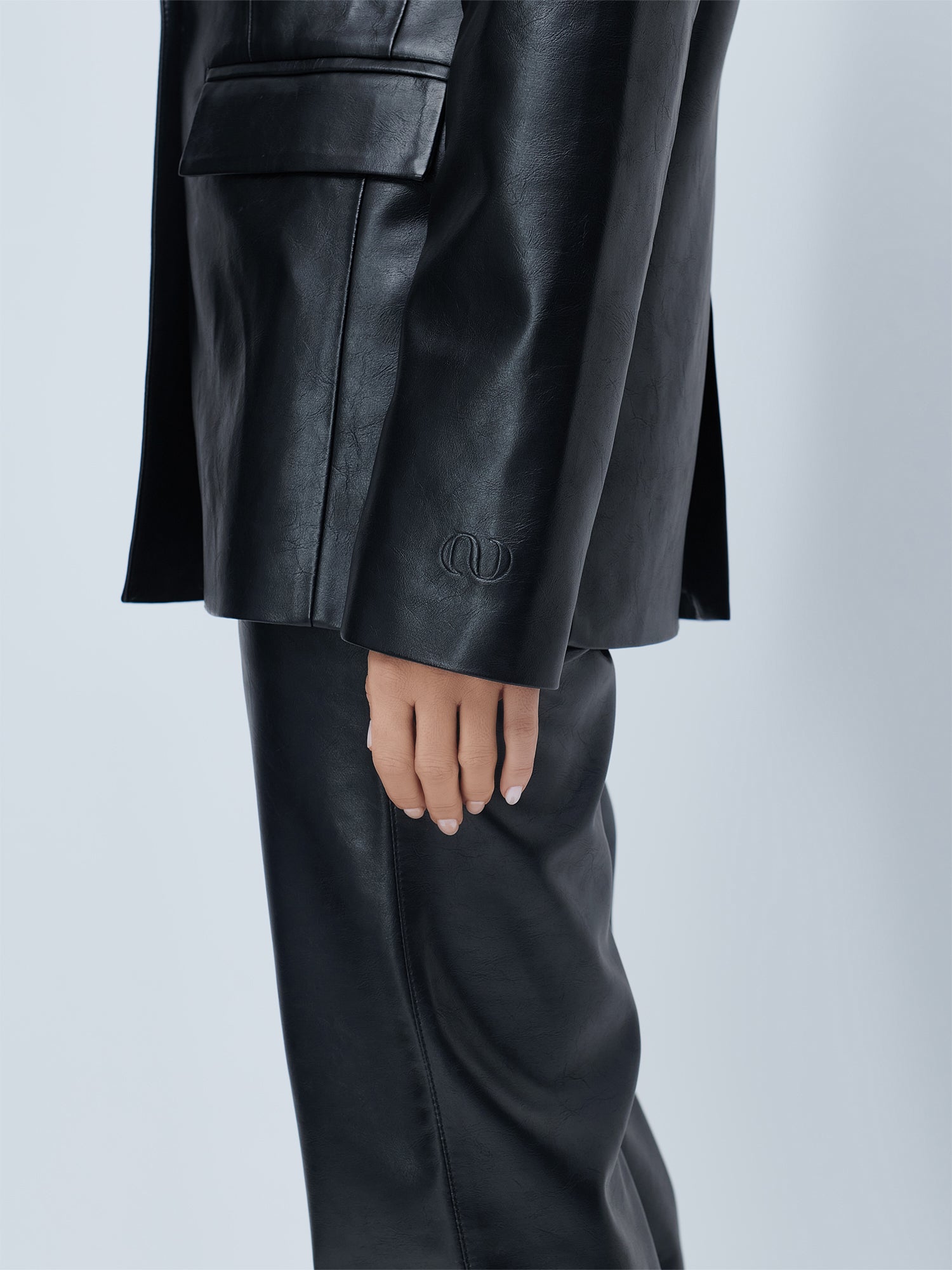 Eine Person trägt den schwarzen „Insa“-Blazer von LeGer by Lena Gercke im Einreiher-Design mit passender Hose, den Arm an der Seite. Der Ärmel weist nahe der Manschette dezente Prägungen auf, die sich von einem schlichten, hellen Hintergrund abheben.