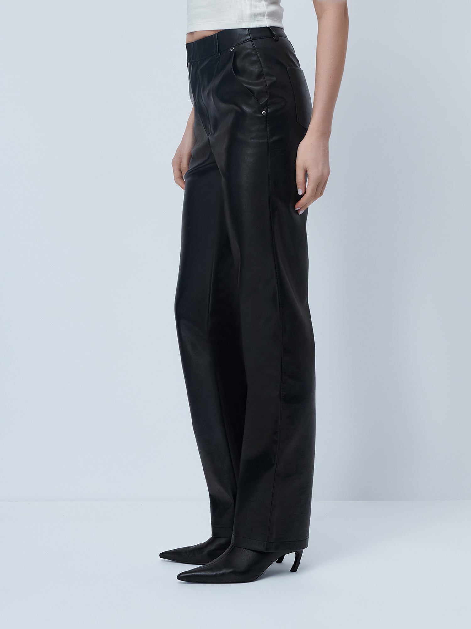 Eine Person, die die Hose „Insa“ von LeGer by Lena Gercke trägt – aus schwarzem Leder mit hoher Bundhöhe und geradem Bein –, kombiniert sie mit schwarzen Stiefeln mit spitzer Zehenpartie und Absatz sowie einem weißen, ärmellosen Oberteil und steht vor einem schlichten, hellen Hintergrund.