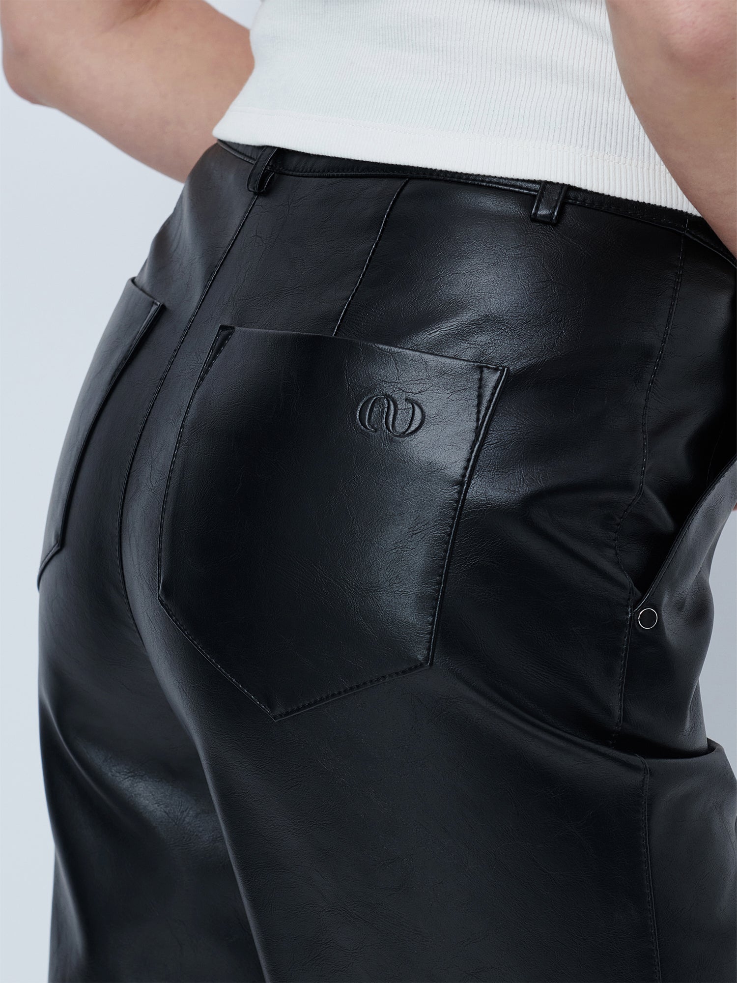 Nahaufnahme einer Person, die die „Insa“-Hose von LeGer by Lena Gercke trägt – eine schwarze Kunstlederhose mit geradem Bein, hohem Bund und sichtbaren Gesäßtaschen (eine davon mit gesticktem Logo), kombiniert mit einem weißen, gerippten Top.