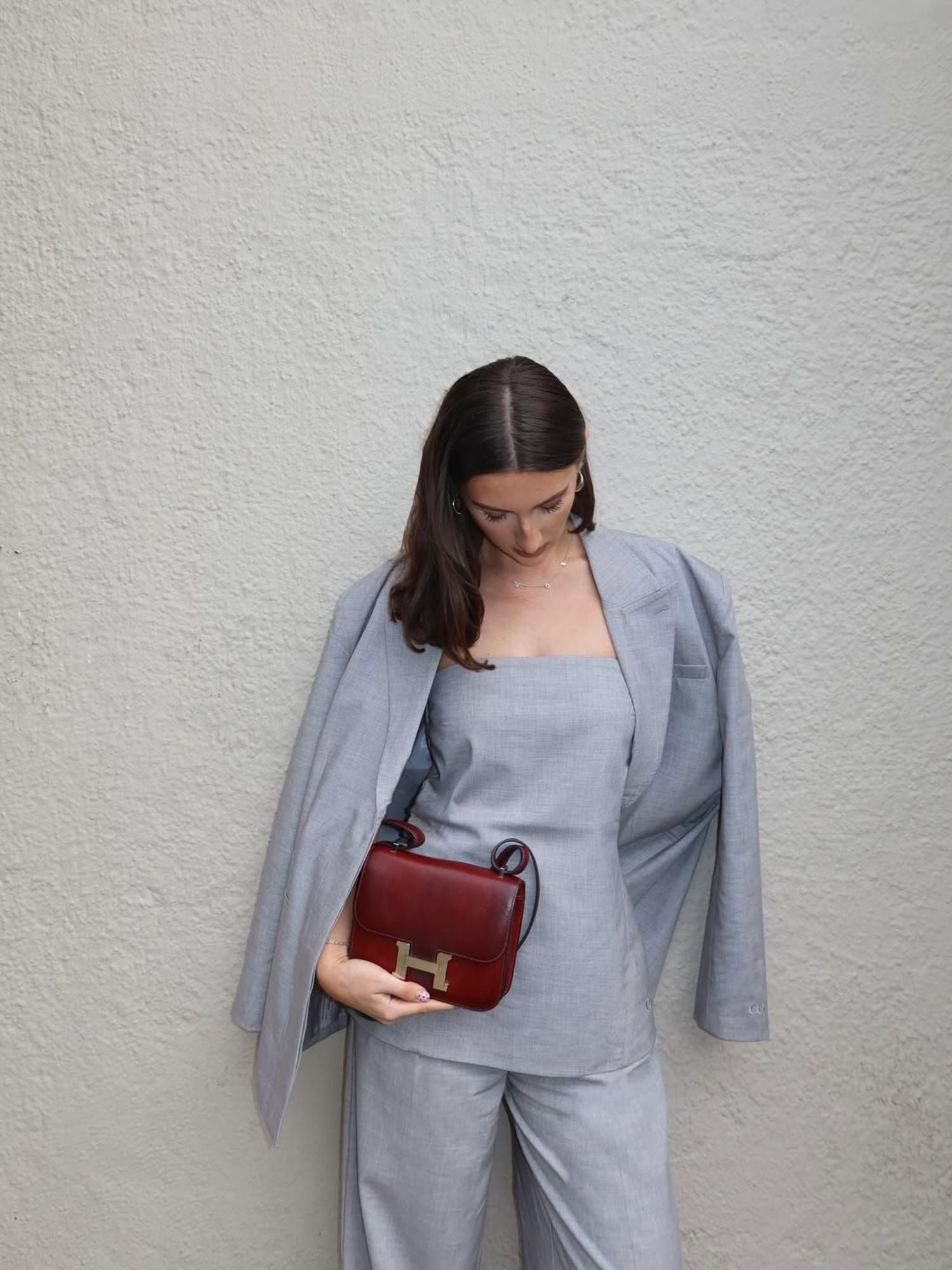 Eine Frau in einem hellgrauen Anzug drapiert ihren Blazer über die Schultern und blickt auf eine kleine dunkelrote Handtasche. Sie trägt das Jale Top von LeGer by Lena Gercke, dessen schulterfreies Design vor einer strukturierten Wand mühelose Eleganz verleiht.