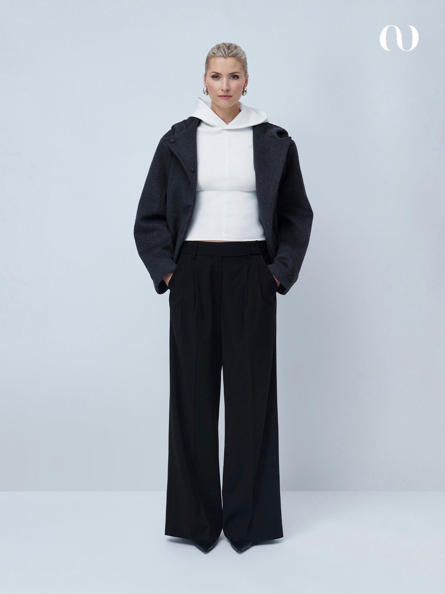 Vor einem hellgrauen Hintergrund trägt eine Frau die Chinara Pants von LeGer by Lena Gercke in lockerer Passform, kombiniert mit weißem Hoodie und dunkler Jacke. Sie hat blonde, zurückgebundene Haare und schaut geradeaus, die Hände in den Taschen.