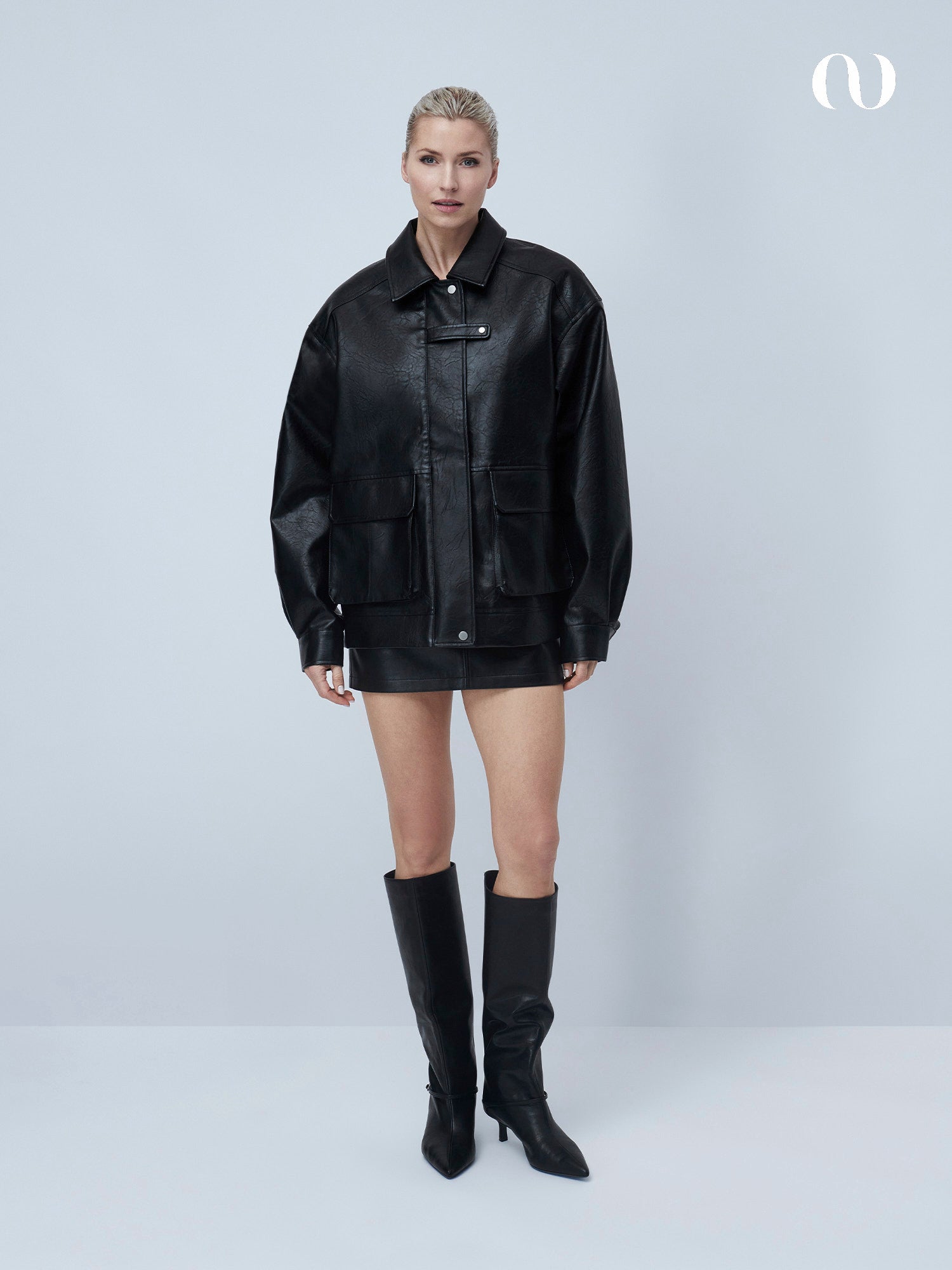 Eine Frau steht vor einem schlichten, hellen Hintergrund und trägt die Davinia Jacket von LeGer by Lena Gercke, gepaart mit einem schwarzen Minirock und kniehohen Stiefeln. Ihr Haar ist hochgesteckt und sie blickt mit neutralem Blick in die Kamera.