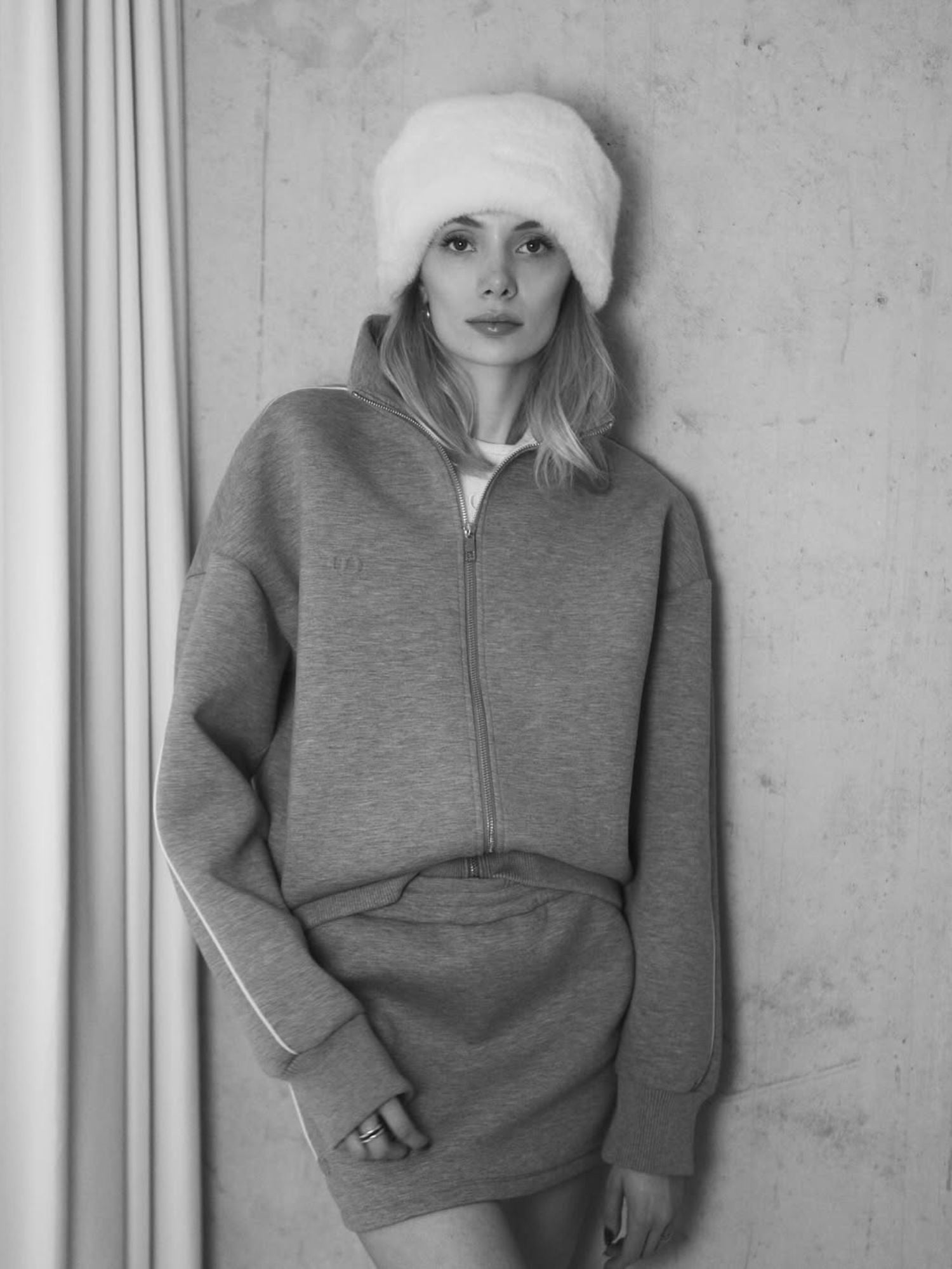 Schwarz-Weiß-Foto einer Frau, die vor einer strukturierten Wand steht. Sie trägt eine wuschelige weiße Mütze, ein Sweatshirt mit Reißverschluss und den Lilou Rock von LeGer by Lena Gercke. Ihr Haar ist offen, ihr Gesichtsausdruck neutral, und eine Hand ruht an ihrer Seite.
