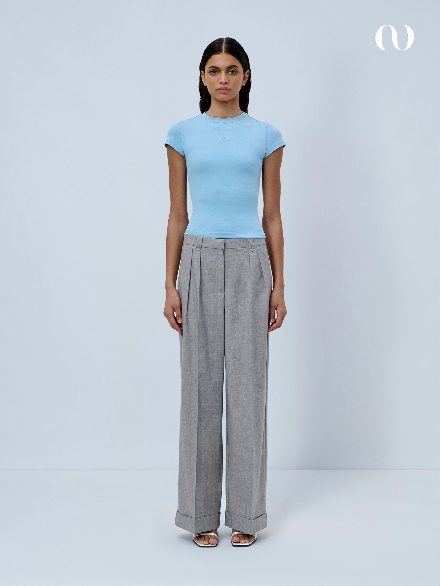 Eine Frau steht vor einem schlichten hellen Hintergrund und trägt ein hellblaues LeGer by Lena Gercke Franka T-Shirt und eine graue Hose mit hoher Taille und weitem Bein. Sie kombiniert den Look mit offenen Absätzen und hat einen neutralen Ausdruck.