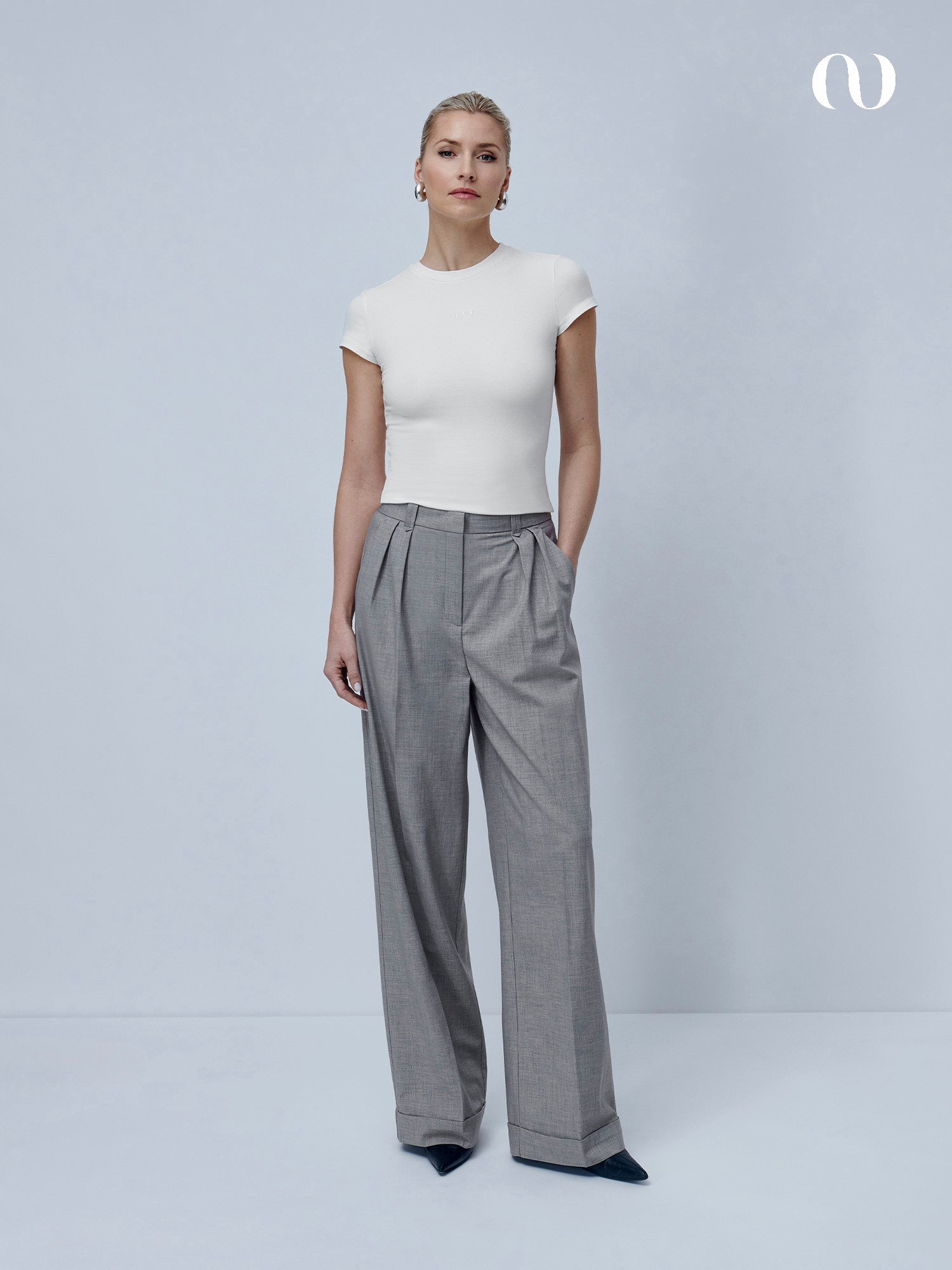 Eine Frau steht vor einem unifarbenen, hellgrauen Hintergrund und trägt das LeGer by Lena Gercke Franka T-Shirt in schmaler Passform mit Rundhalsausschnitt, gepaart mit einer hochtaillierten, grauen, plissierten Hose mit weitem Bein und spitzen schwarzen Schuhen. Ihr Haar ist zurückgesteckt und sie hat einen neutralen Ausdruck.