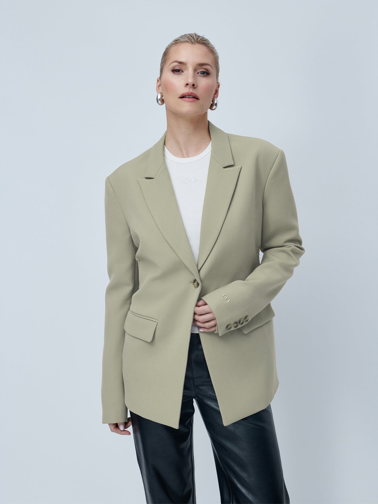 Eine Person mit kurzen blonden Haaren und Creolen trägt den Blazer der LeGer by Lena Gercke Line mit langen Ärmeln und entspannter Passform über einem weißen Hemd und einer schwarzen Lederhose und steht mit selbstbewusstem Gesichtsausdruck vor einem schlichten hellen Hintergrund.