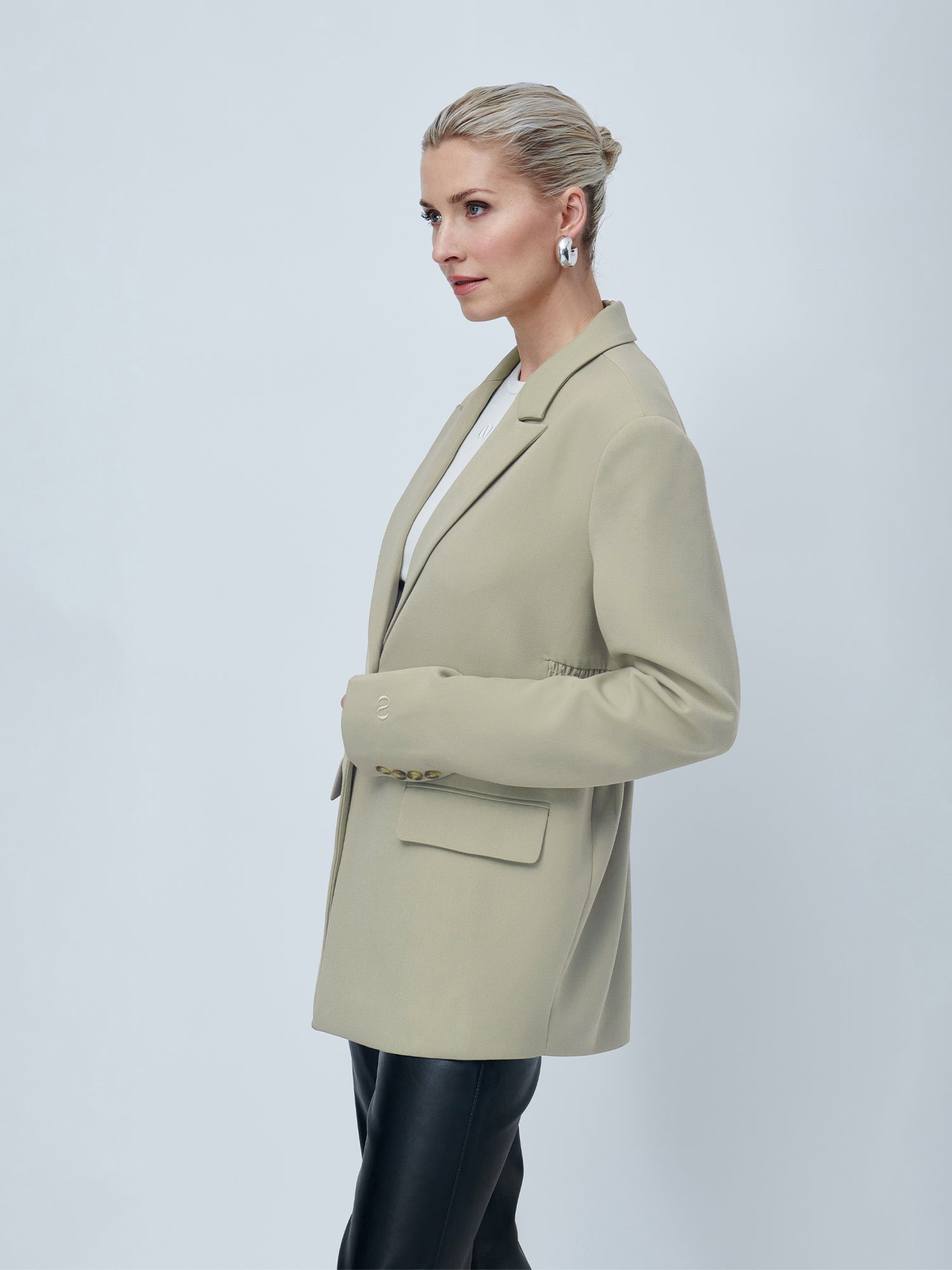 Eine Frau mit blondem Haar zu einem ordentlichen Dutt steht im Profil vor einem schlichten hellgrauen Hintergrund und trägt den LeGer by Lena Gercke Line Blazer in Hellbeige über einem weißen Oberteil, einer schwarzen Lederhose und silbernen Ohrringen.