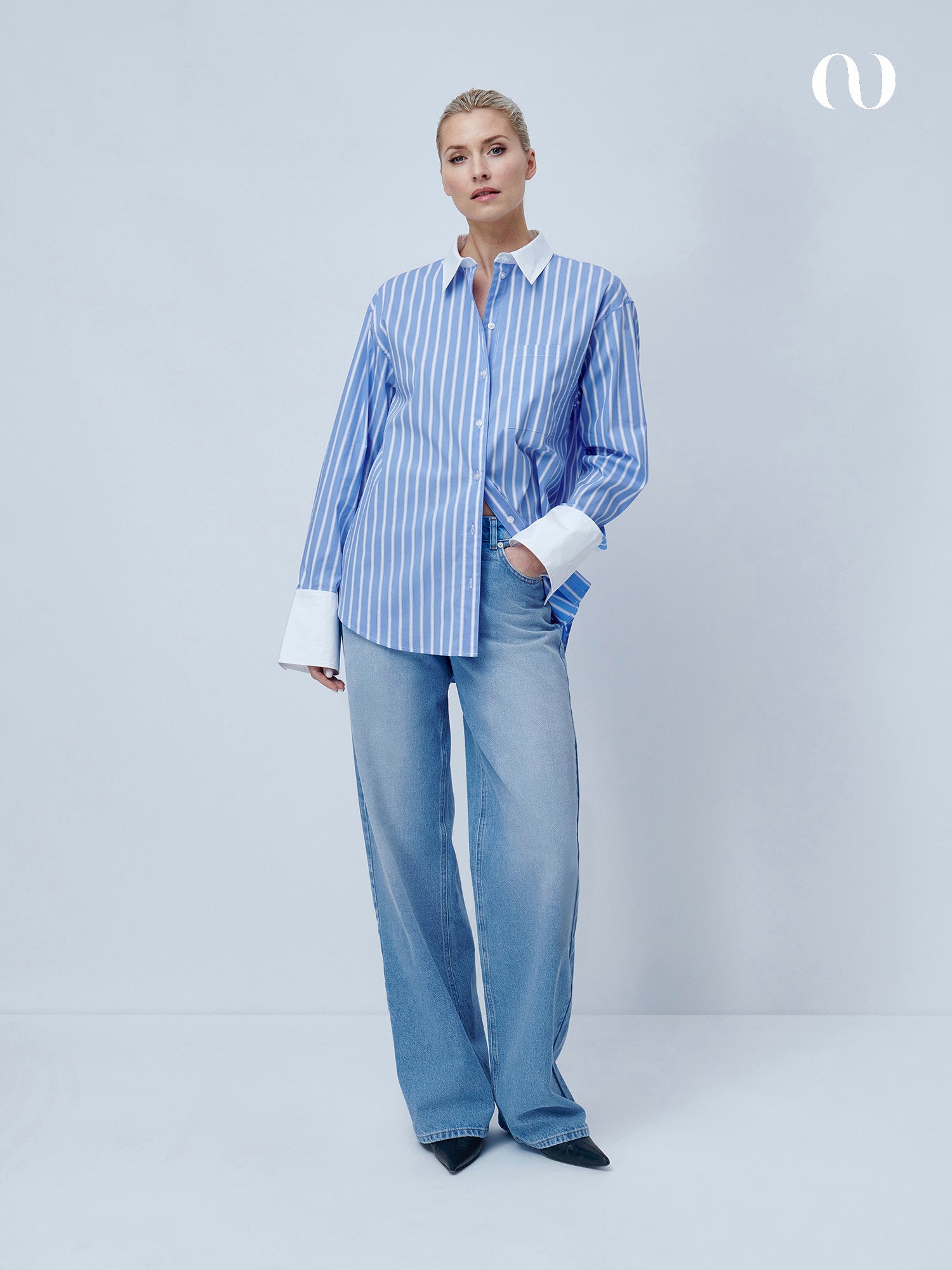 Eine Frau mit kurzen blonden Haaren posiert vor einem hellen Hintergrund und trägt die LeGer by Lena Gercke Jarina Blouse mit blauen und weißen Streifen, langen Ärmeln und lockerer Passform, gepaart mit hellblauen Jeans mit weitem Bein und schwarzen spitzen Schuhen.