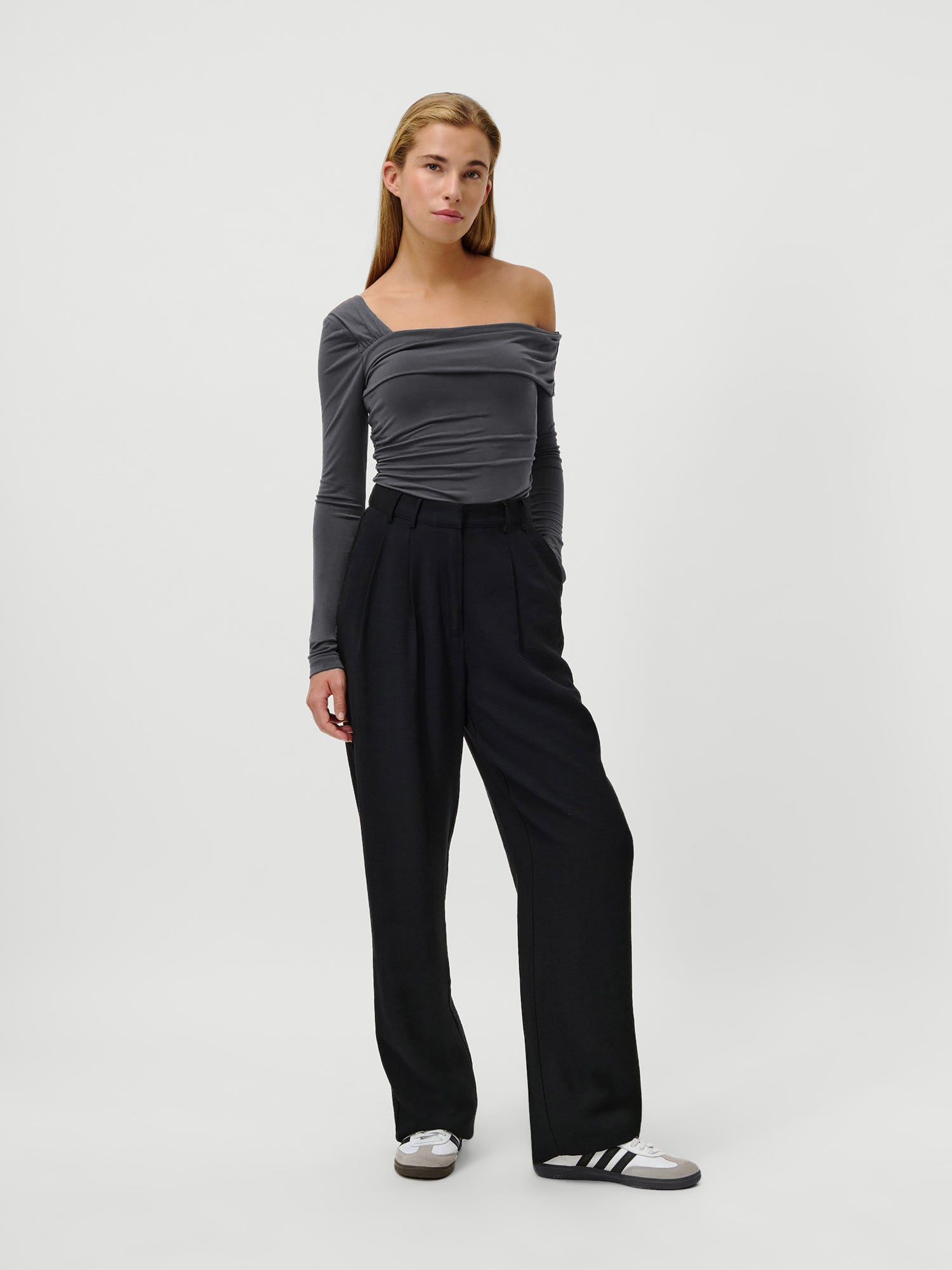 Eine Frau steht vor einem weißen Hintergrund und trägt das LeGer by Lena Gercke Charleen Longsleeve - ein schmal geschnittenes, schulterfreies graues Oberteil mit extra langen Ärmeln - kombiniert mit einer schwarzen Hose mit hoher Taille und weißen Adidas-Sneakern und blickt ruhig in die Kamera.