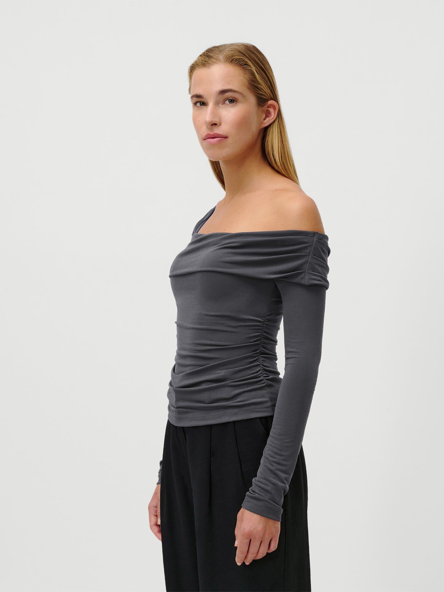 Eine Frau mit glattem blondem Haar trägt das Charleen Longsleeve von LeGer by Lena Gercke - ein schmal geschnittenes, schulterfreies graues Oberteil mit extralangen Ärmeln - in Kombination mit einer schwarzen Hose. Sie steht vor einem schlichten weißen Hintergrund und blickt leicht nach links.