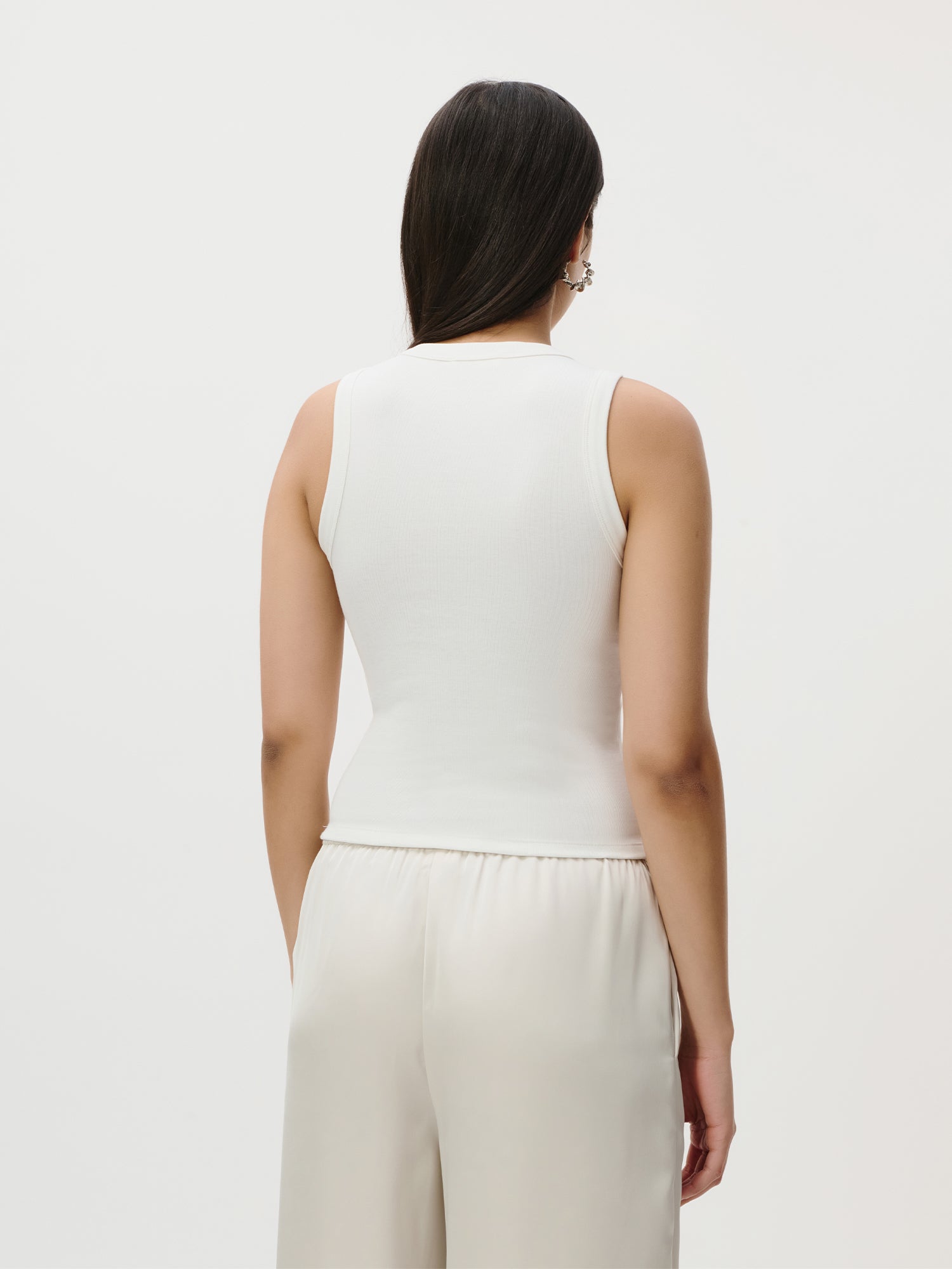 Eine Person mit langen dunklen Haaren steht mit dem Rücken zur Kamera vor einem schlichten hellen Hintergrund und trägt das ärmellose „Ria“-Top von LeGer by Lena Gercke sowie eine lockere weiße Hose aus einer minimalistischen Capsule Wardrobe.