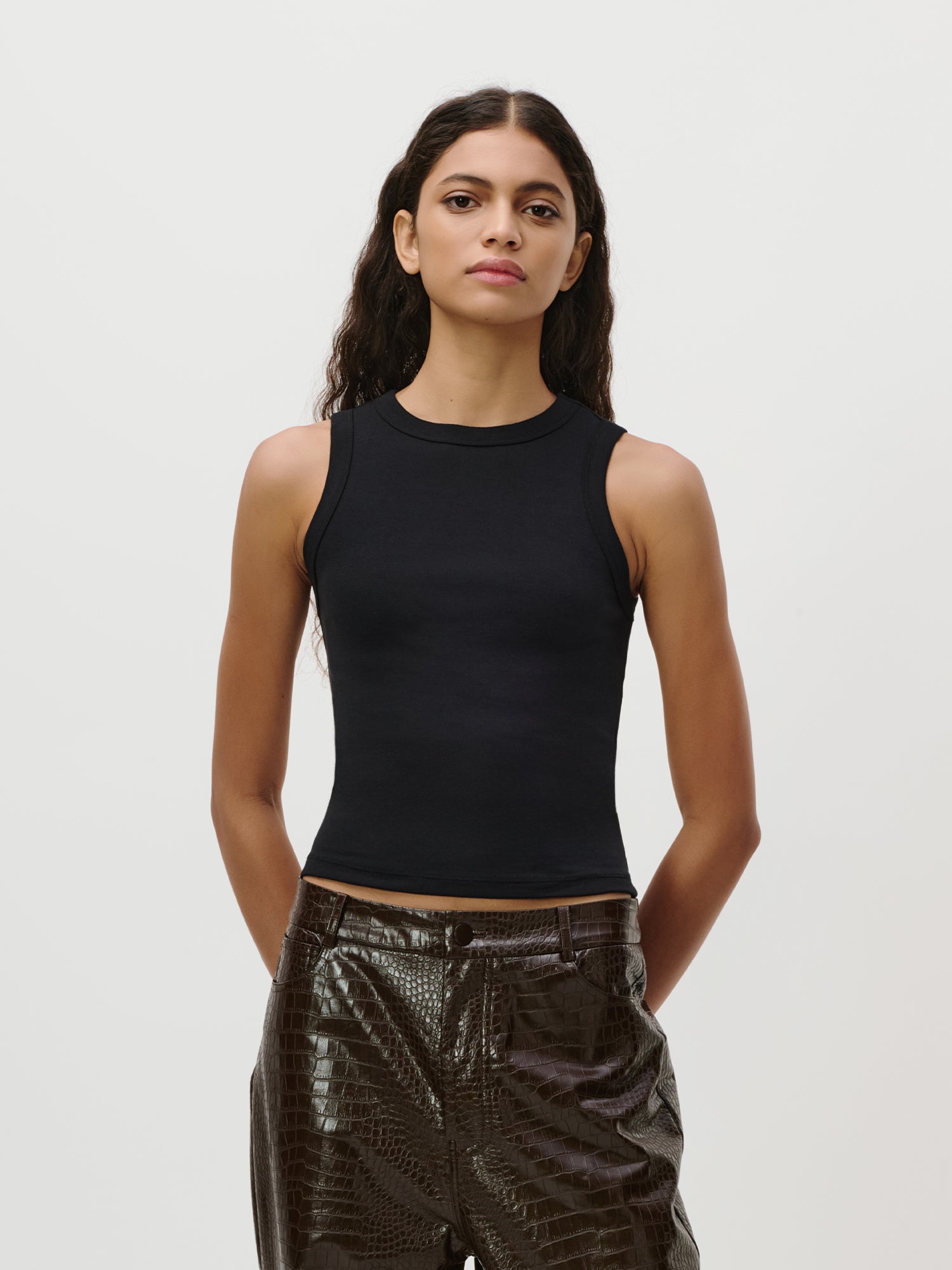 Eine Person mit langen, welligen dunklen Haaren trägt das „Ria“-Top von LeGer by Lena Gercke – ein ärmelloses Oberteil –, kombiniert mit glänzenden, strukturierten dunklen Hosen mit dezenter Rippenstruktur, und steht mit den Händen in den Taschen vor einem schlichten weißen Hintergrund.