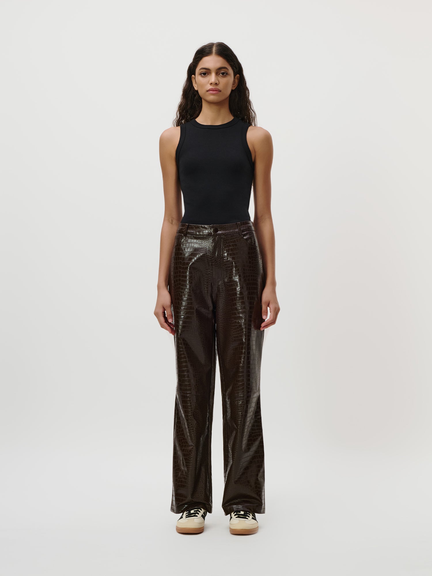 Eine Person steht mit dem Blick nach vorne vor einem schlichten weißen Hintergrund und trägt das schwarze „Ria“-Top von LeGer by Lena Gercke mit dezenter Ripp-Struktur, eine glänzende, dunkelbraune Hose mit Struktur sowie beigefarbene Sneaker – ein vielseitiger Look, der sich perfekt für jede Capsule Wardrobe eignet.