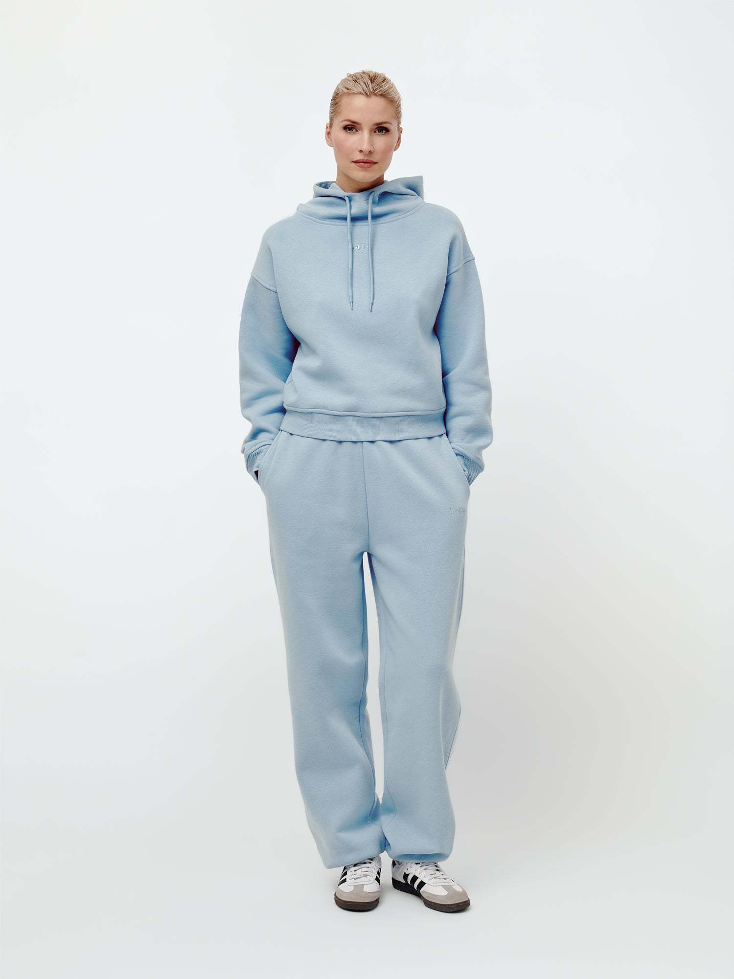 Eine Person steht frontal im Bild und trägt einen hellblauen LeGer by Lena Gercke Hoodie im „Karima“-Stil mit langen Ärmeln und lockerer Passform sowie eine dazu passende Sweathose in regulärer Länge; die Hände stecken lässig in den Taschen. Sie hat kurzes blondes Haar und trägt außerdem schwarz-weiße Sneaker. Der Hintergrund ist schlicht weiß.