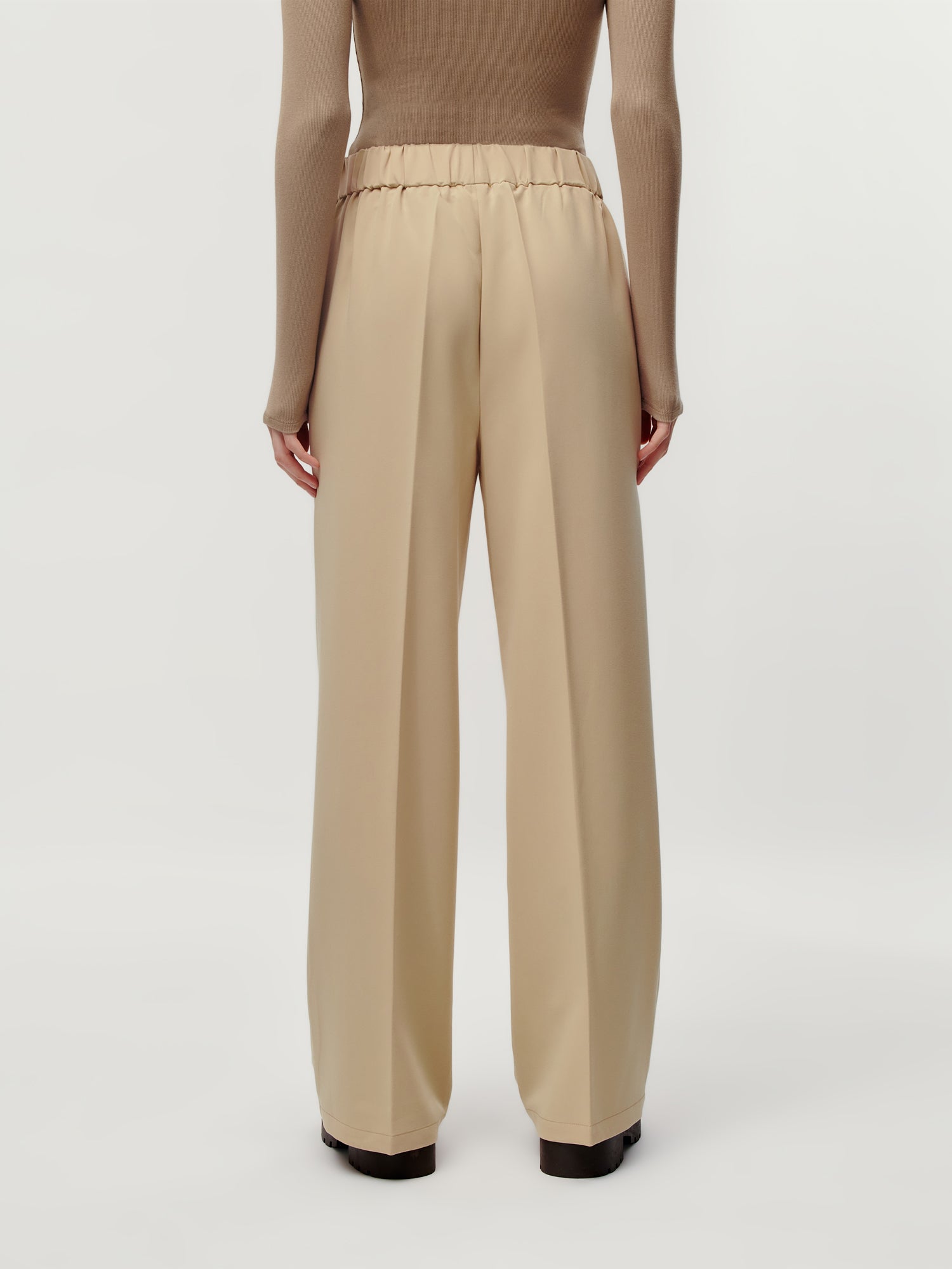 Eine Person ist von hinten zu sehen, die die Allie Pants von LeGer by Lena Gercke in Beige, Größe 36, mit einer Maxi-Länge für einen eleganten Look trägt, gepaart mit einem passenden langärmeligen Oberteil vor einem schlichten hellen Hintergrund.