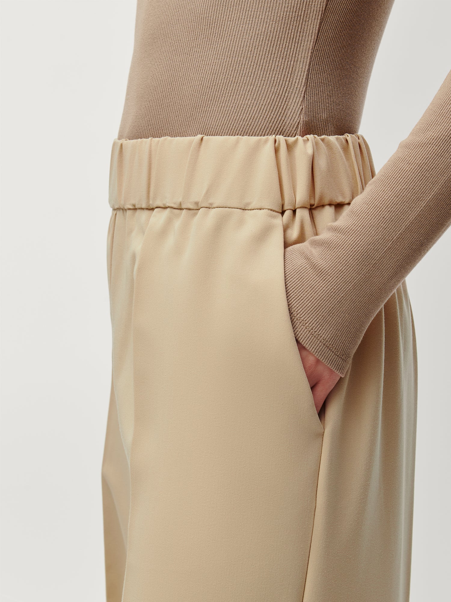 Eine Person in LeGer by Lena Gercke Allie Pants, beige high-waisted Größe 36 mit elastischem Bund, und einem gerippten hellbraunen langärmeligen Oberteil steht mit einer Hand in der Tasche vor einem einfarbigen Hintergrund.