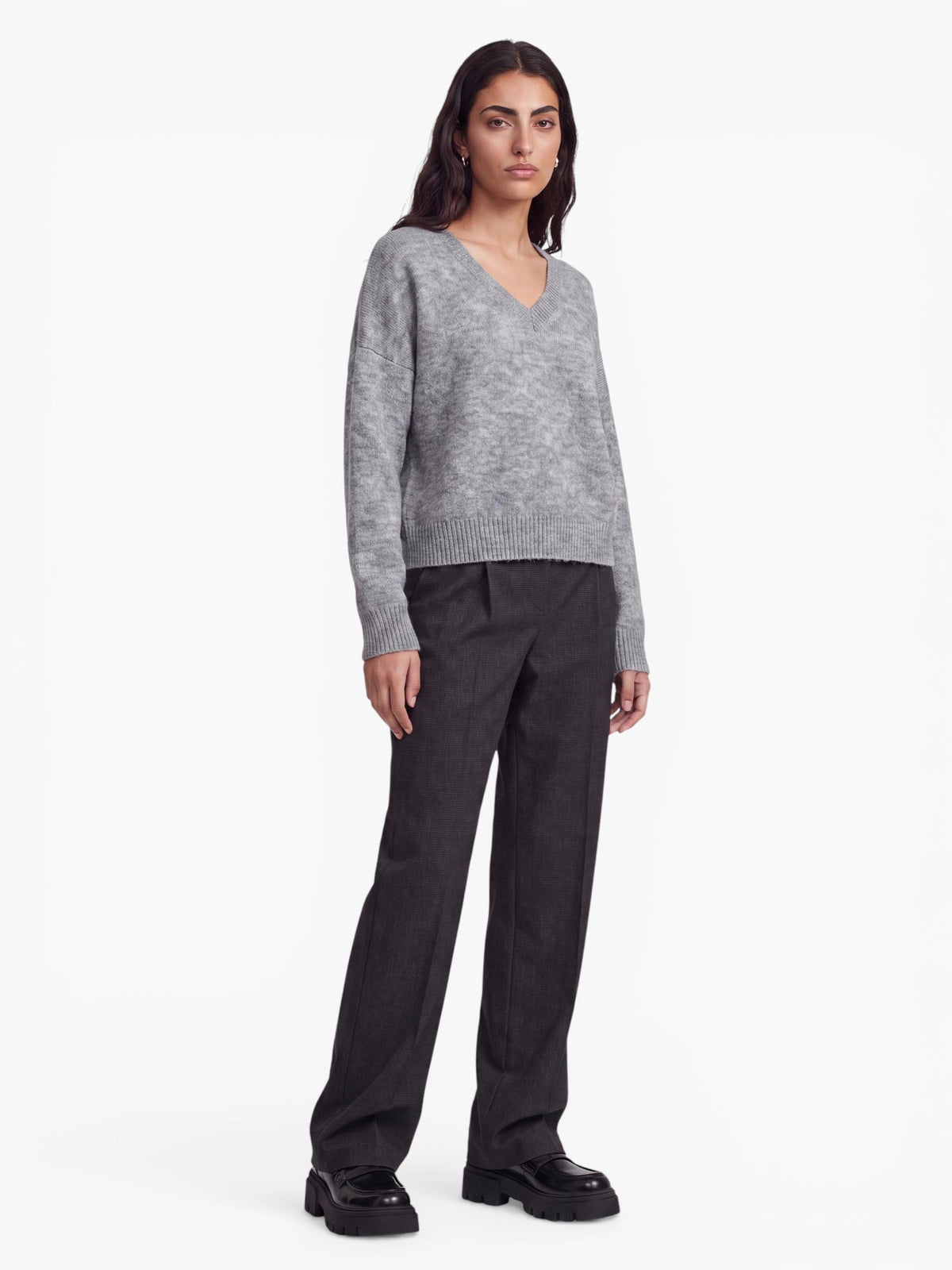 Vor einem weißen Hintergrund steht eine Frau, die den übergroßen grauen „Halina“-Pullover von LeGer by Lena Gercke trägt, dazu eine dunkelgraue Hose mit weitem Bein und schwarze Plateauschuhe.
