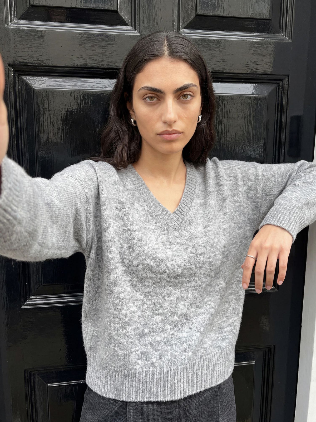 Eine Person mit welligem, dunklem Haar und hellen Augen steht vor einer schwarzen Tür. Sie trägt den übergroßen grauen „Halina“-Pullover von LeGer by Lena Gercke mit V-Ausschnitt und eine dunkle Hose; ein Arm ist angewinkelt, während der andere aus dem Bild hinausragt.