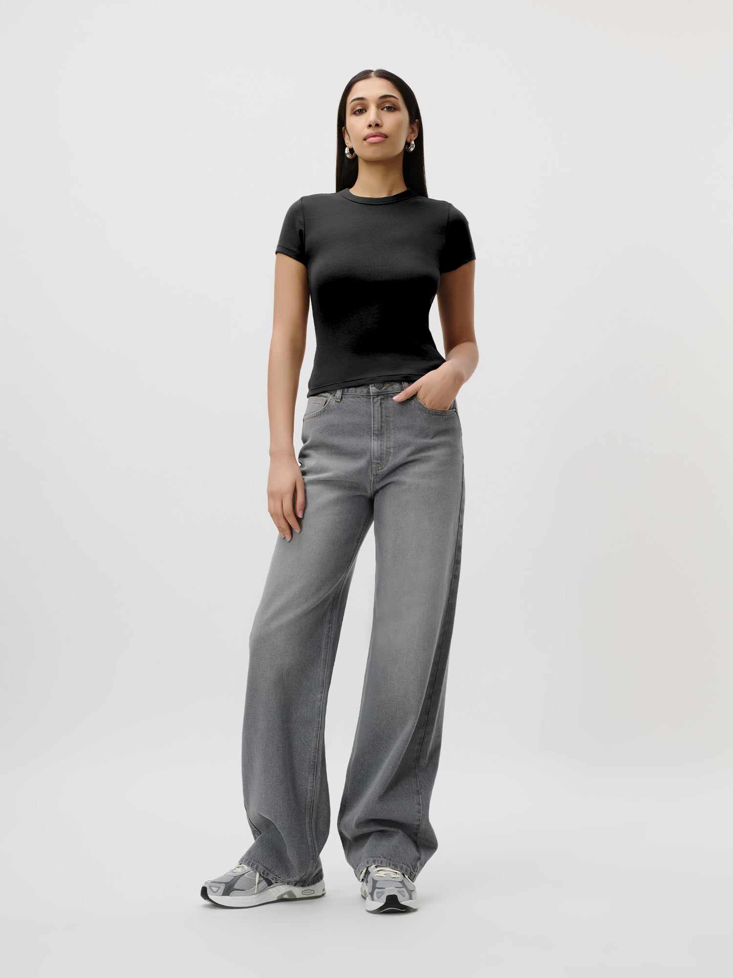 Eine Frau steht vor einem schlichten Hintergrund und trägt ein eng anliegendes schwarzes „Ria“-T-Shirt von LeGer by Lena Gercke, dazu eine graue Jeans mit weitem Bein und Sneaker. Eine Hand steckt in ihrer Hosentasche, während sie selbstbewusst nach vorne blickt.