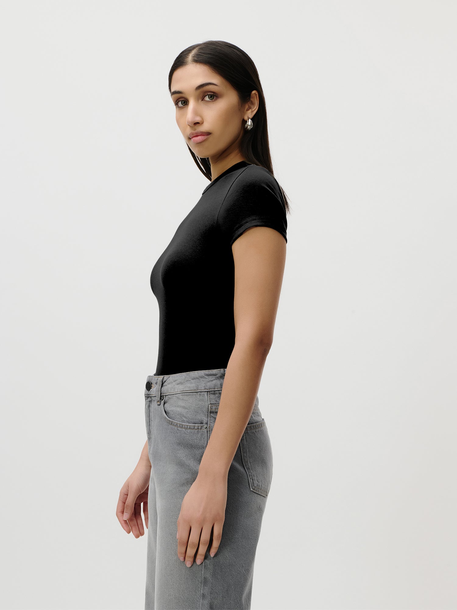 Eine Person mit glattem, dunklem Haar trägt das schmal geschnittene „Ria“-T-Shirt von LeGer by Lena Gercke in Schwarz, dazu eine graue Jeans. Sie steht seitlich vor einem schlichten weißen Hintergrund und blickt leicht in Richtung Kamera.