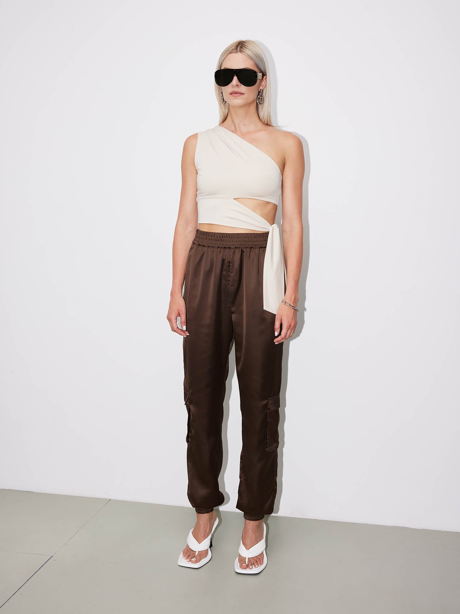 Eine Frau mit langen blonden Haaren trägt eine schwarze Sonnenbrille, das beigefarbene Leia Top von LeGer by Lena Gercke (schmaler Schnitt), braune Satin-Jogginghosen, weiße Sandaletten mit Absatz und große Ohrringe und steht vor einer schlichten weißen Wand.