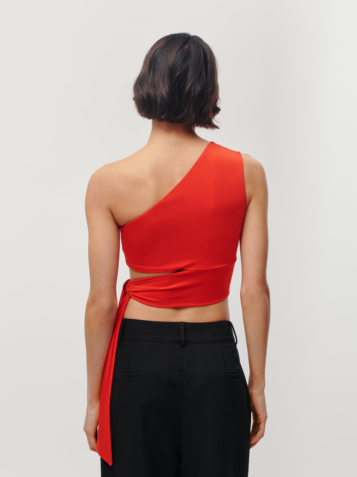Von hinten ist eine Person mit kurzen, dunklen Haaren zu sehen, die das leuchtend rote Leia-Top von LeGer by Lena Gercke trägt – ein ärmelloses One-Shoulder-Design mit Wickeldetail und kurzer, schmaler Passform – kombiniert mit einer schwarzen Hose vor einem schlichten hellen Hintergrund.