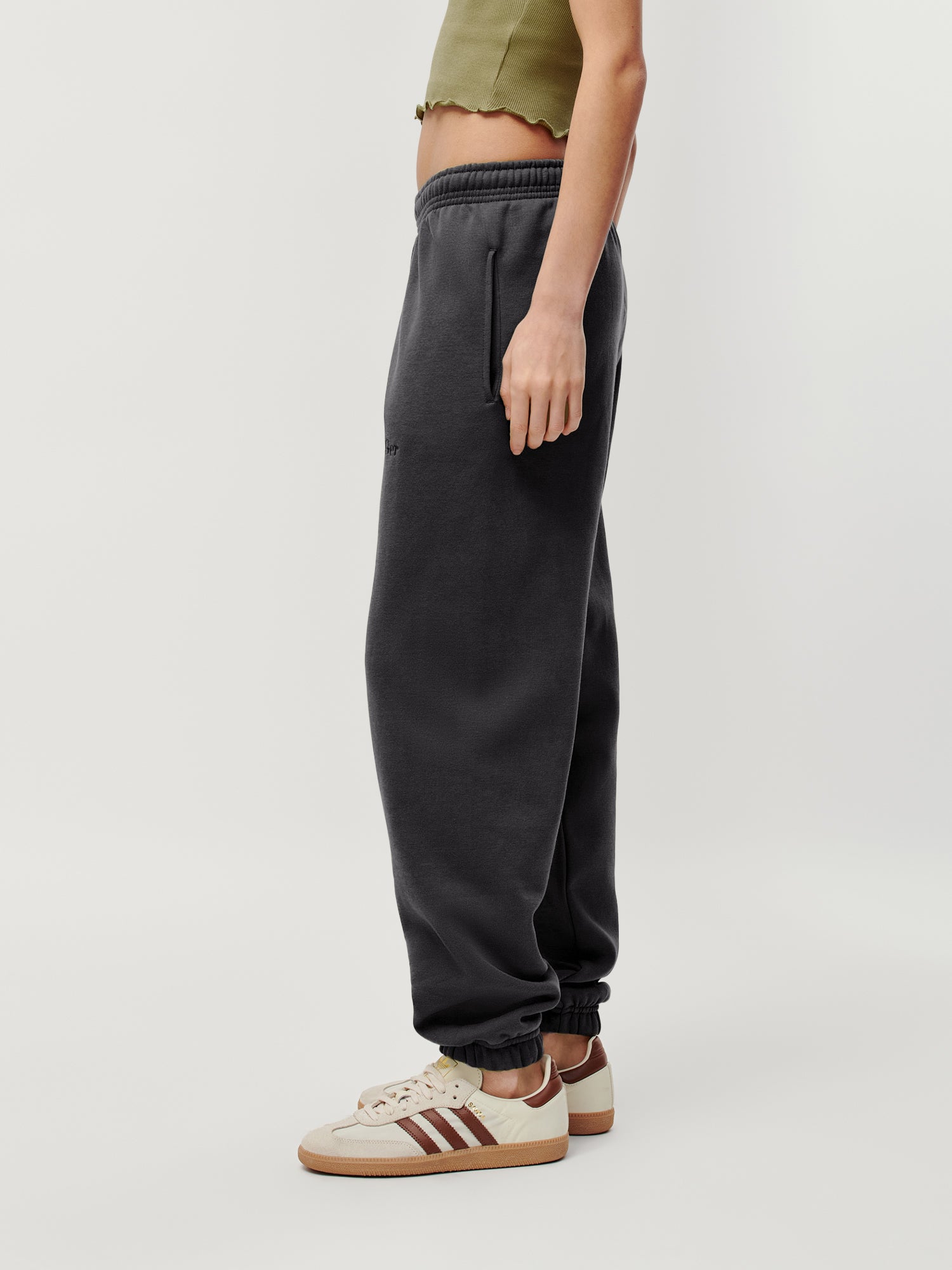 Eine Person steht seitlich und trägt die Ruby Pants von LeGer by Lena Gercke in Dunkelgrau, gepaart mit einem olivgrünen Oberteil und beigen Turnschuhen mit braunen Streifen vor einem schlichten weißen Hintergrund.