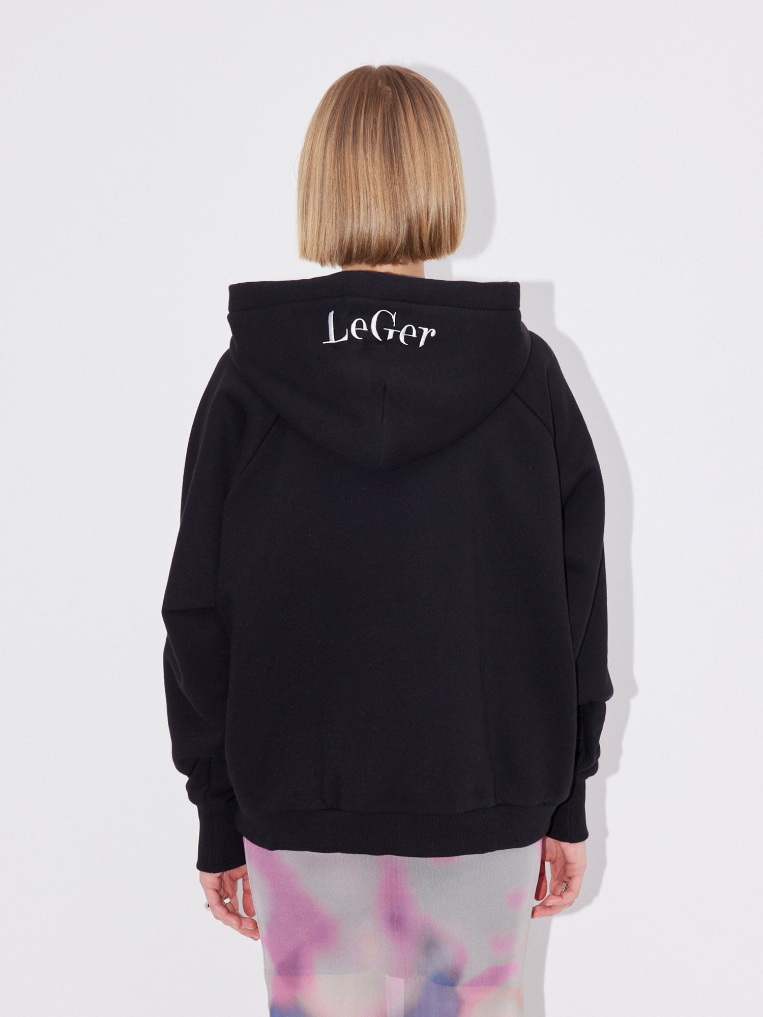 Eine Person mit kurzen blonden Haaren blickt von sich weg und trägt einen schwarzen LeGer by Lena Gercke Hayley Hoodie mit "LeGer" in weißer Schrift auf der Rückseite der Kapuze und einen Rock mit pastellfarbenem Muster. Der Hayley Hoodie hat eine normale Länge und Passform.