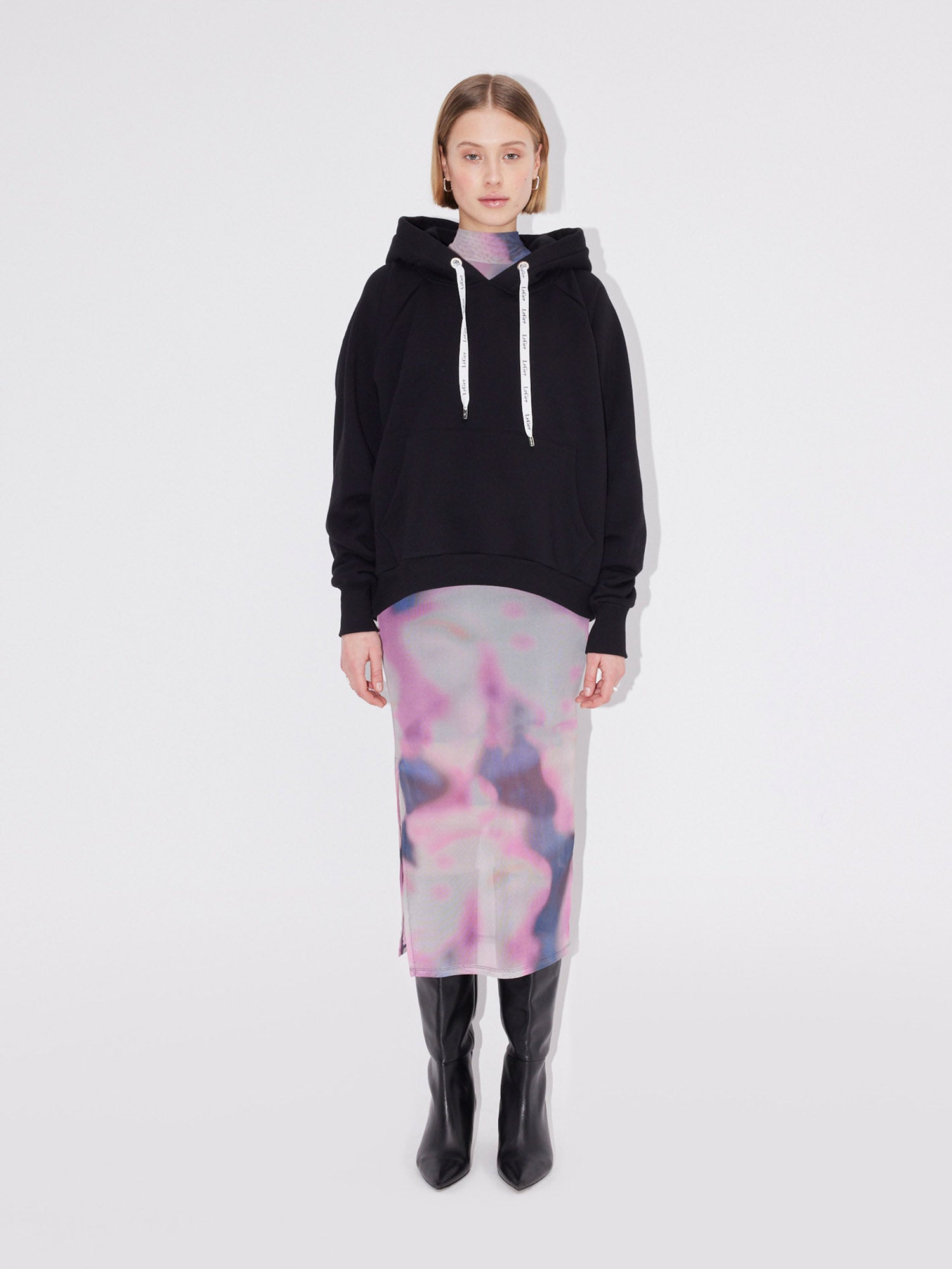 Eine Frau steht vor einem schlichten weißen Hintergrund und trägt den schwarzen Hayley Hoodie von LeGer by Lena Gercke über einem abstrakt gemusterten Kleid in Pastellrosa und Lila, dazu kniehohe schwarze Stiefel. Sie schaut geradeaus und hat die Arme an den Seiten verschränkt.