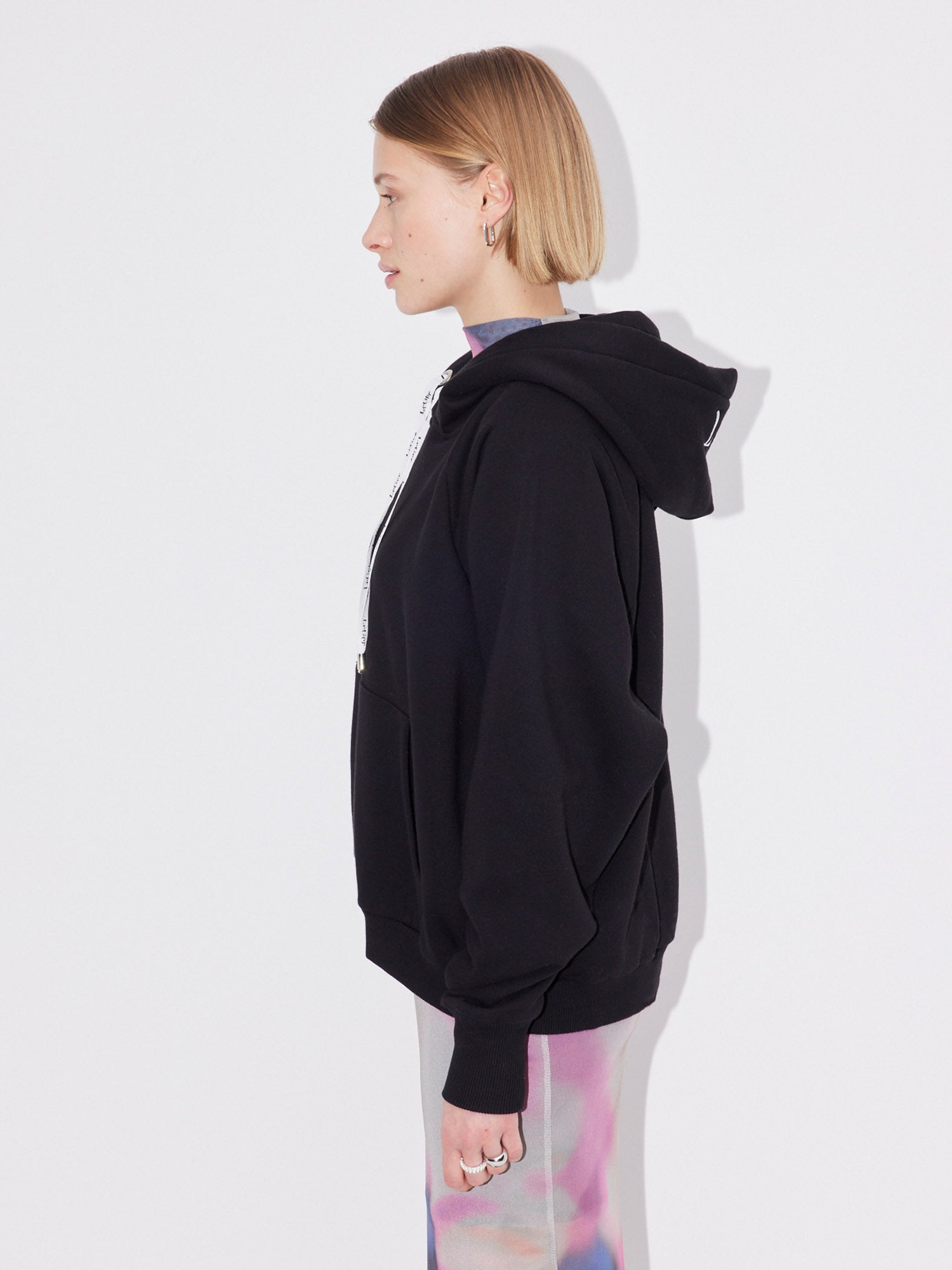 Eine Person mit kurzen, hellbraunen Haaren steht im Profil vor einem weißen Hintergrund und trägt den LeGer by Lena Gercke Hayley Hoodie in Schwarz und eine bunte, pastellfarbene Hose in normaler Länge.