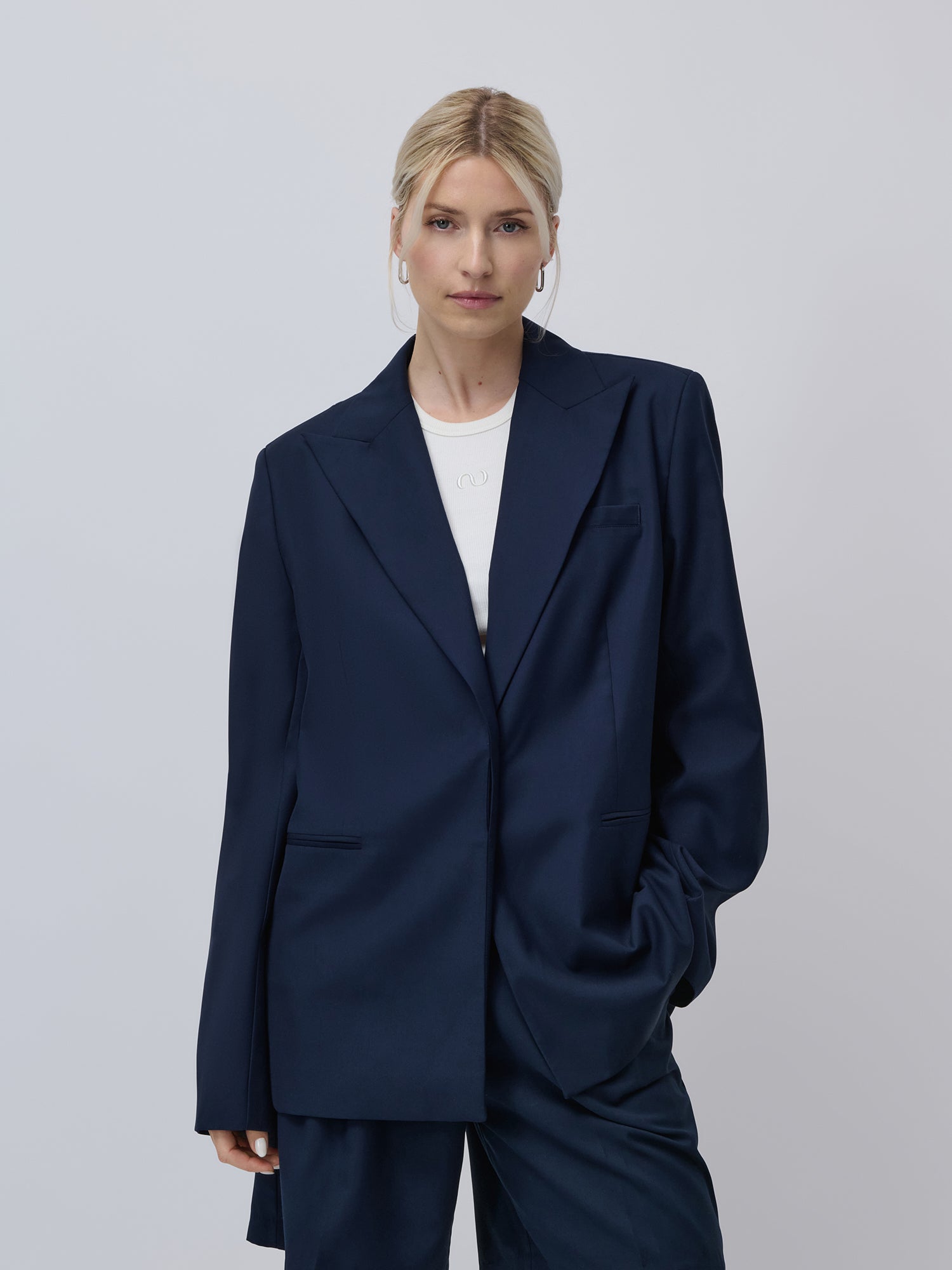 Eine Frau mit zurückgebundenen blonden Haaren trägt das marineblaue Dita Set von LeGer by Lena Gercke - Blazer und passende Hose über einem weißen Oberteil. Sie steht vor einem hellgrauen Hintergrund und hat eine Hand in ihrer Tasche. Set Preisvorteil verfügbar.