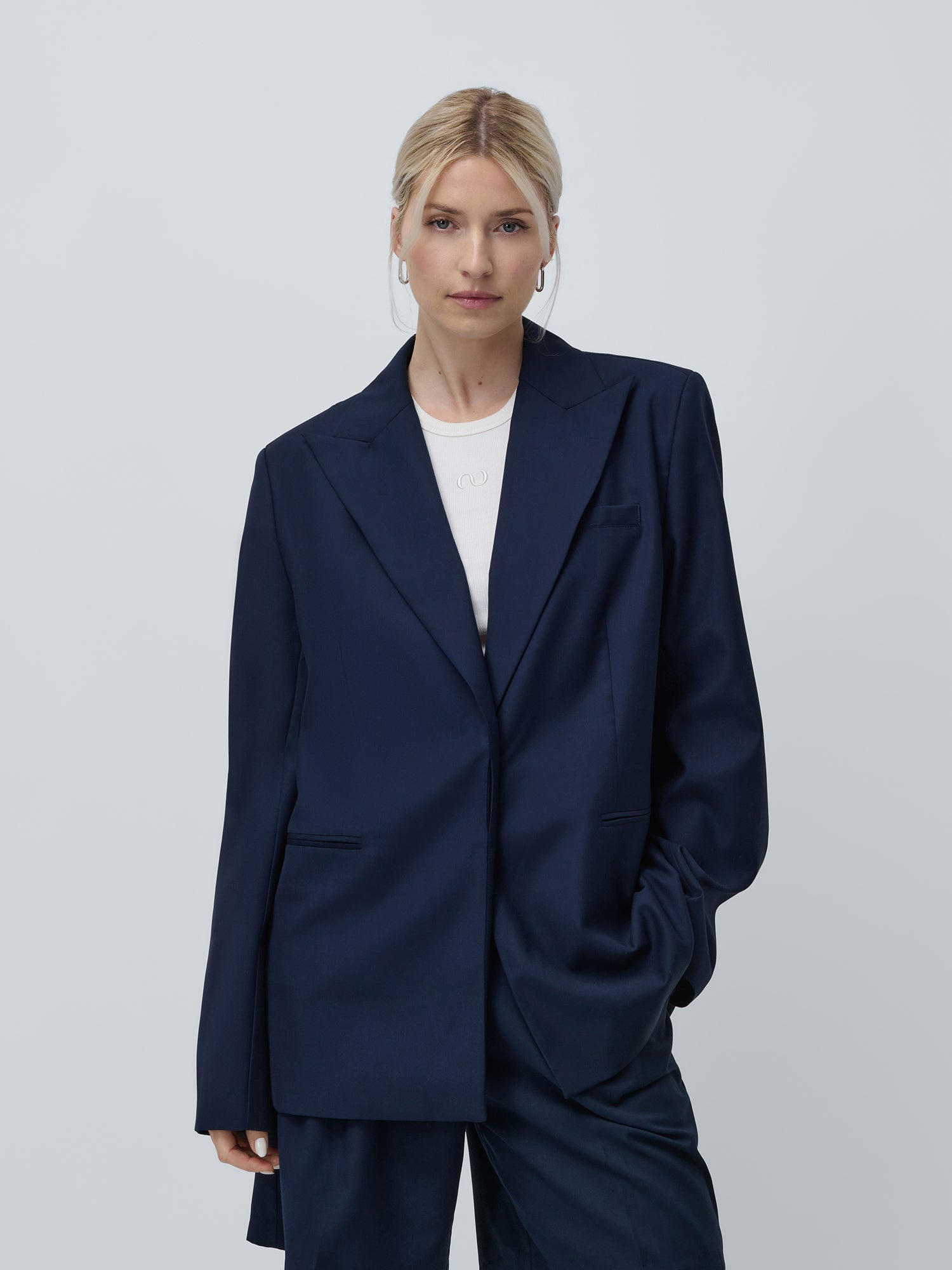 Eine Person mit blonden Haaren trägt den Dita Blazer von LeGer by Lena Gercke über einem frischen weißen Hemd und steht selbstbewusst mit einer Hand in der Tasche vor einem schlichten Hintergrund.