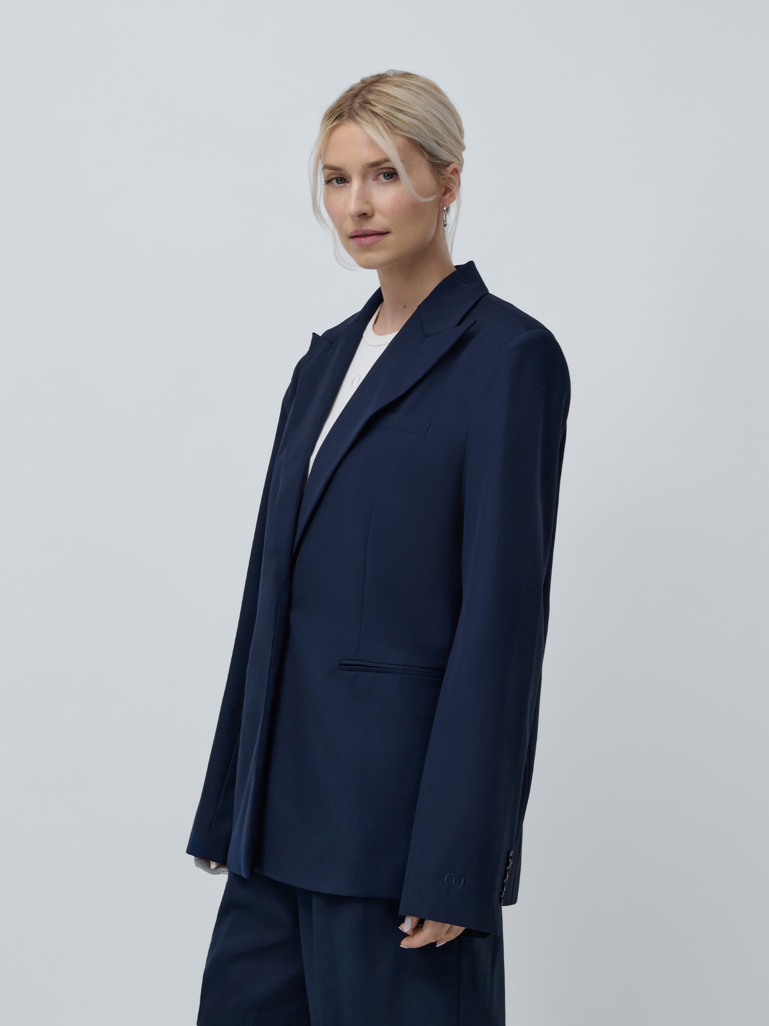 Eine blonde Person trägt den LeGer by Lena Gercke Dita Blazer, eine dunkelblaue Jacke mit stylischem Spitzrevers, über einem weißen Hemd auf hellgrauem Hintergrund. Sie hat einen neutralen Gesichtsausdruck und ihre Hände ruhen an den Seiten.