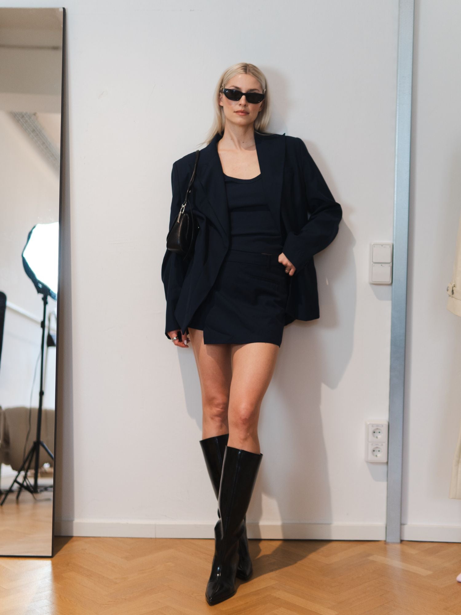 Eine Person trägt den LeGer by Lena Gercke Dita Blazer in navy mit passendem Rock, schwarze kniehohe Stiefel, eine Sonnenbrille und eine schwarze Umhängetasche. Sie posiert selbstbewusst in einem Raum vor einem großen Spiegel an einer weißen Wand.