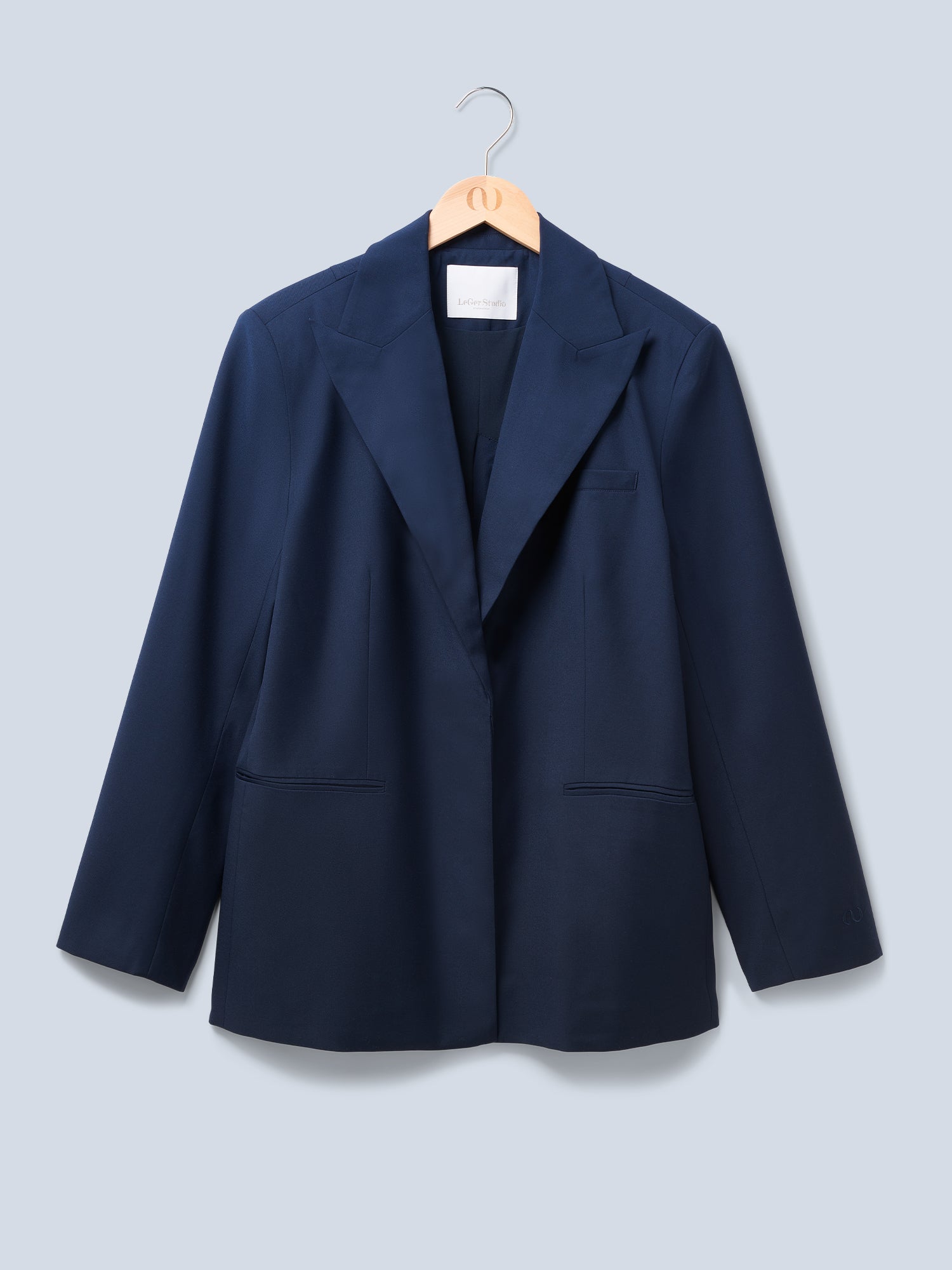 Der Dita Blazer von LeGer by Lena Gercke ist ein marineblauer, locker sitzender Blazer mit Spitzrevers, zwei Vordertaschen und einer Brusttasche, der auf einem Holzbügel vor einem hellgrauen Hintergrund gezeigt wird.