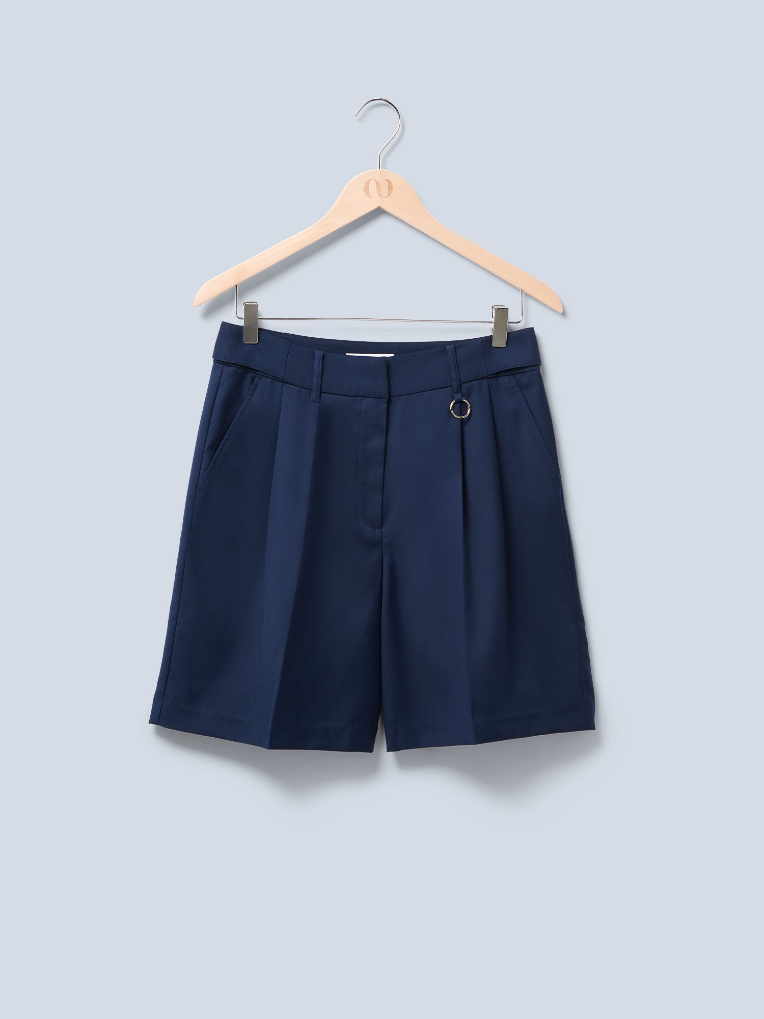 Die Dita Shorts von LeGer by Lena Gercke sind marineblaue, taillierte Shorts mit mittlerer Leibhöhe und Bügelfalten. Sie verfügen über Gürtelschlaufen und einen dekorativen Metallring und hängen elegant auf einem Holzbügel vor hellblauem Hintergrund.
