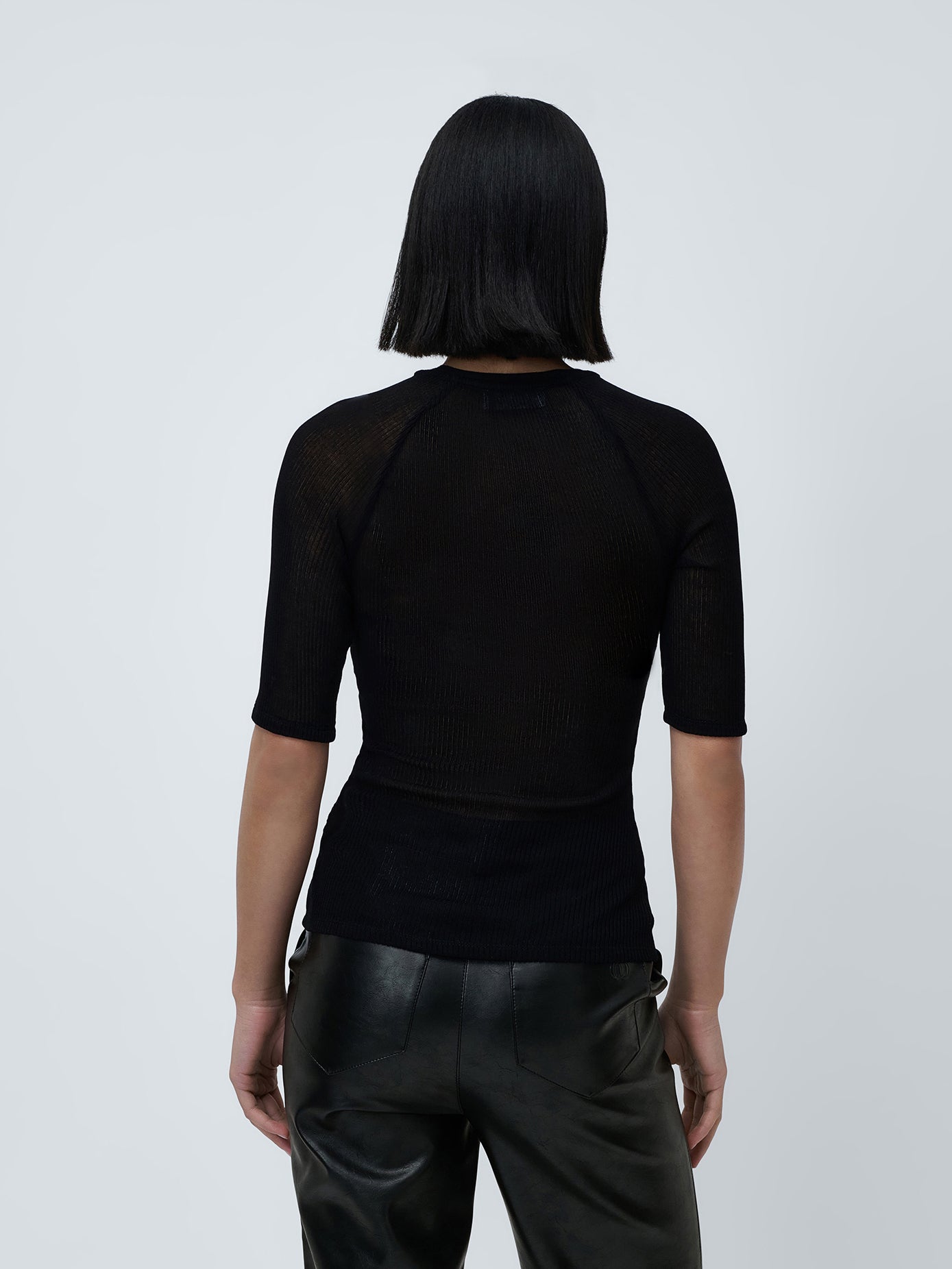 Eine Person mit glattem, schulterlangem, schwarzem Haar ist von hinten zu sehen und präsentiert einen stylischen Look. Sie trägt ein schwarzes Abby T-Shirt von LeGer by Lena Gercke mit halblangen Ärmeln und eine schwarze Lederhose vor einem schlichten hellgrauen Hintergrund.