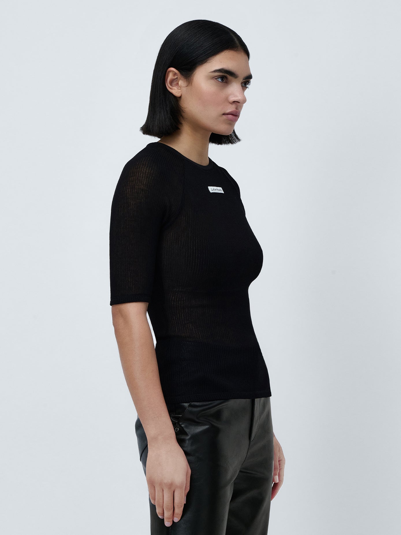 Eine Person mit schulterlangem, dunklem Haar trägt ein Abby-T-Shirt von LeGer by Lena Gercke und eine schwarze Hose. Sie steht vor einem schlichten weißen Hintergrund und blickt mit mühelos stilvollem Aussehen zur Seite.