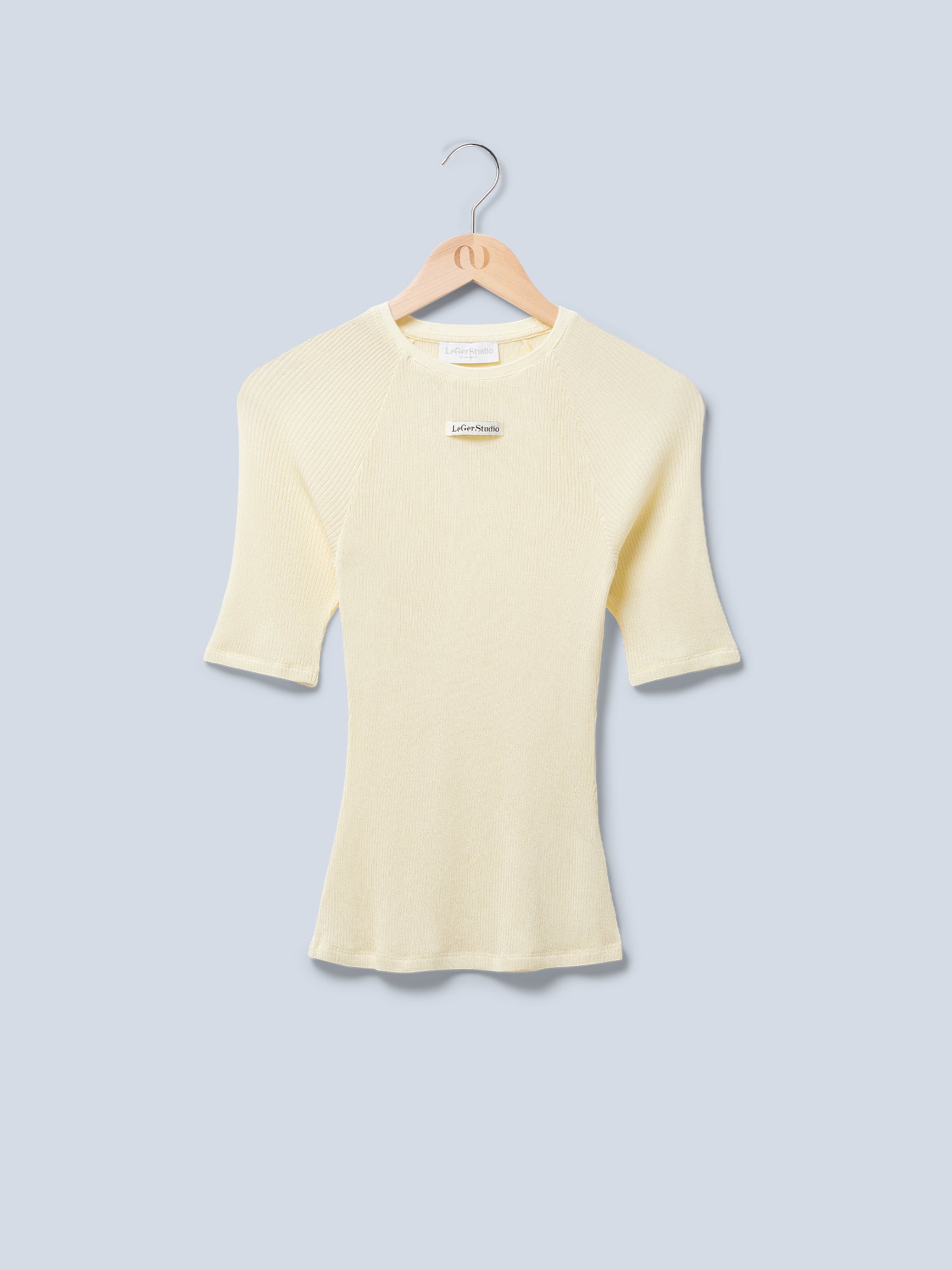 Das Abby T-Shirt von LeGer by Lena Gercke, ein hellgelber Slim Fit, kurzärmliger Rippenpullover mit Rundhalsausschnitt, hängt auf einem Holzbügel vor hellblauem Hintergrund. In der Nähe des Ausschnitts befindet sich ein kleines weißes Etikett mit Text.