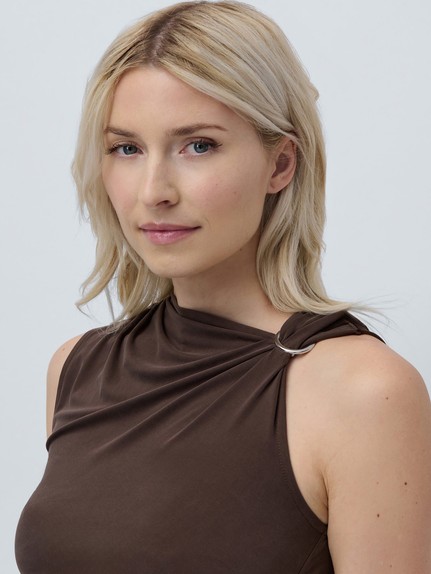 Eine Frau mit hellblondem Haar und heller Haut trägt das Dominique Top von LeGer by Lena Gercke, ein eng anliegendes, ärmelloses Top mit drapiertem, asymmetrischem Ausschnitt und Metallring-Detail an der Schulter. Sie lächelt leicht vor einem schlichten hellen Hintergrund.