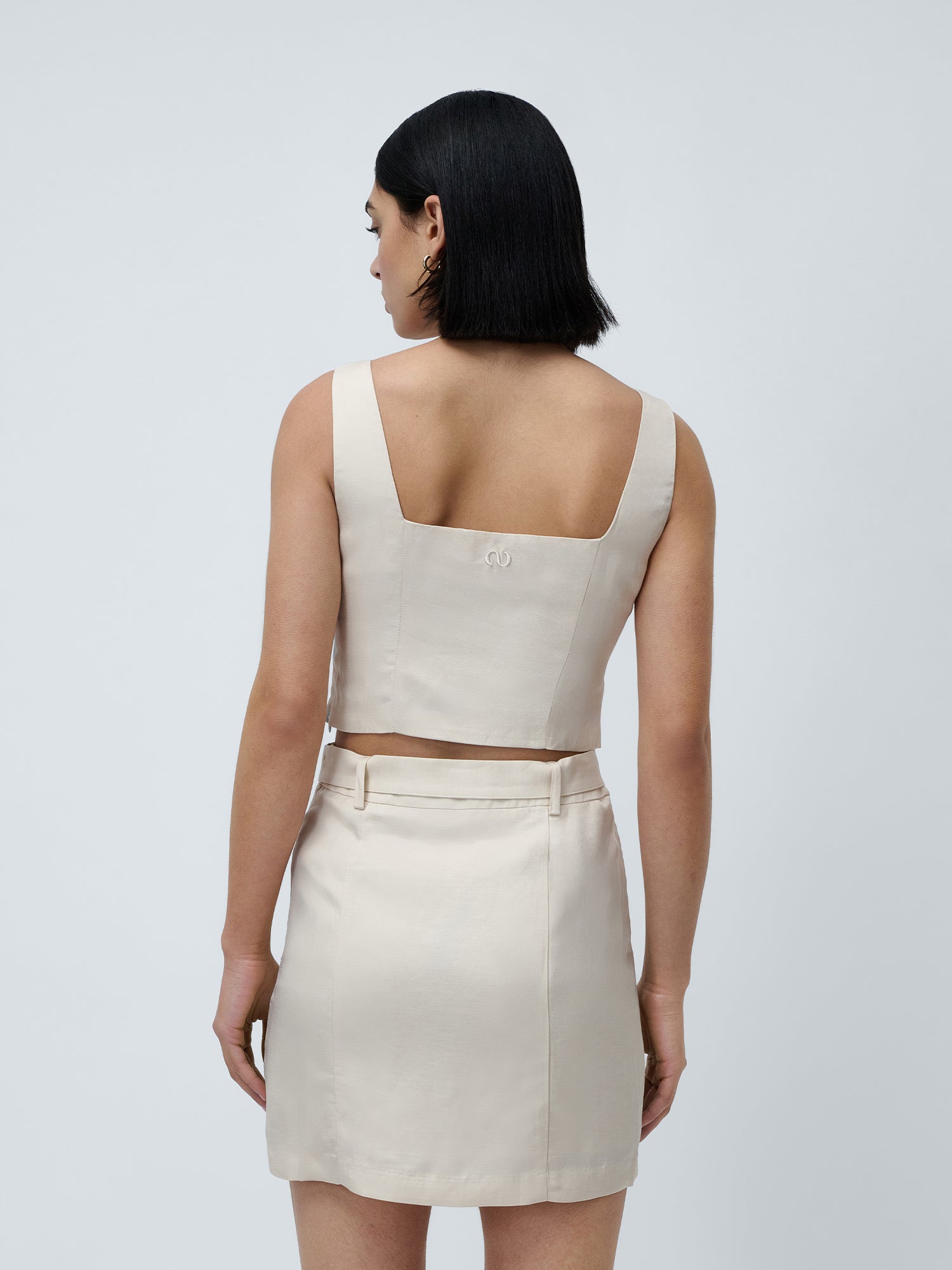 Eine Frau mit kinnlangem, glattem, dunklem Haar ist von hinten zu sehen. Sie trägt das Top „Majella“ von LeGer by Lena Gercke und einen dazu passenden Rock mit hoher Taille – ein schicker Sommerlook vor grauem Hintergrund.